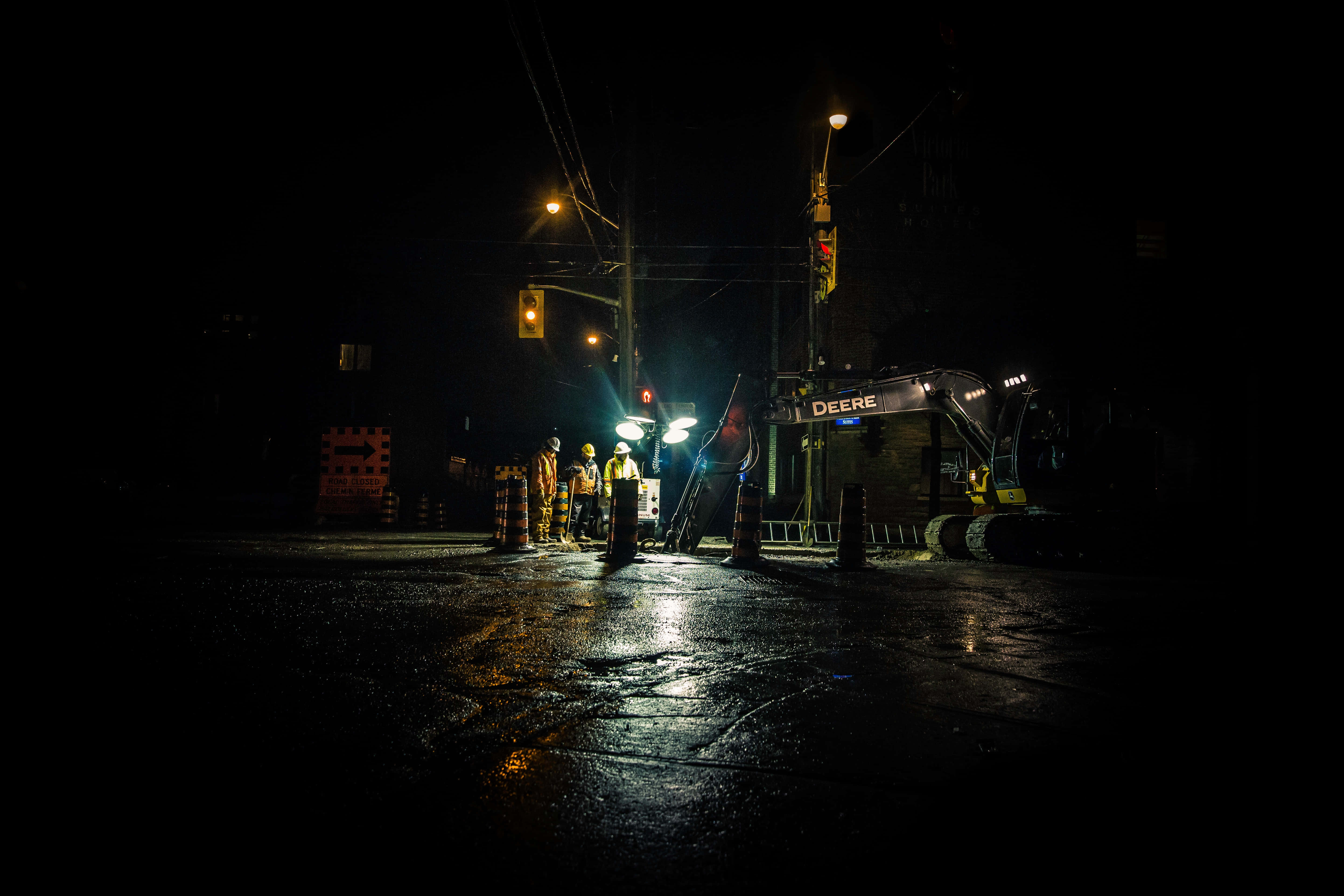 Night Time 桌布 壁紙 — 夜間建築工人被明亮燈光照亮，站在迪爾挖掘機旁邊，位於潮濕的瀝青街道上。