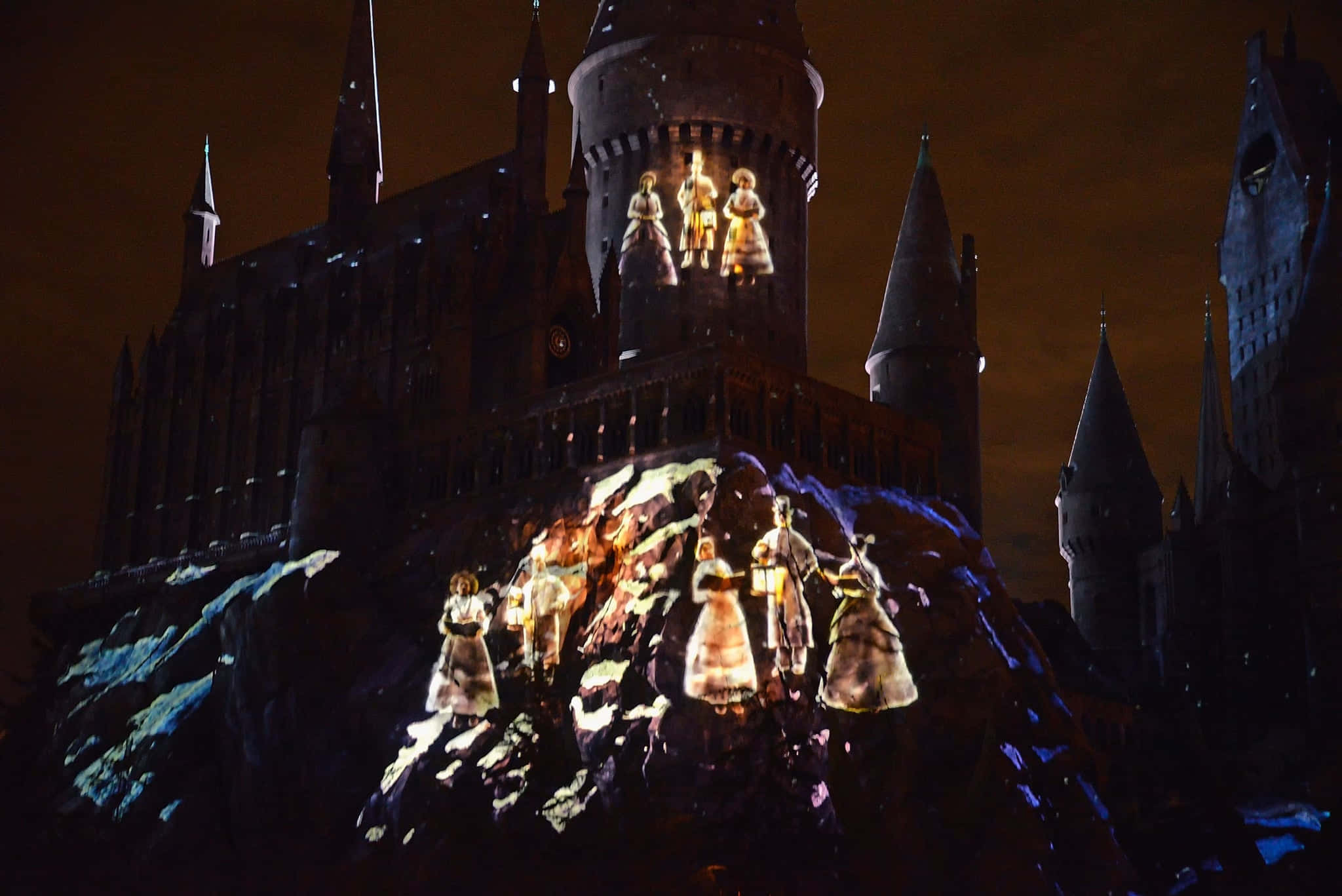 Christmas At Hogwarts 桌布 壁紙 — 夜間投射霍格華茲城堡，動畫角色在黑暗天空下的石牆上發光。