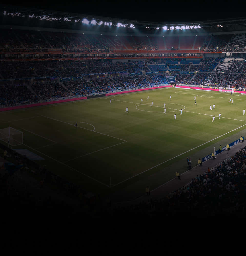 足球 桌布 壁紙 — 夜間明亮照明的足球場鳥瞰圖，球場上有球員，看台座無虛席，聚光燈下。