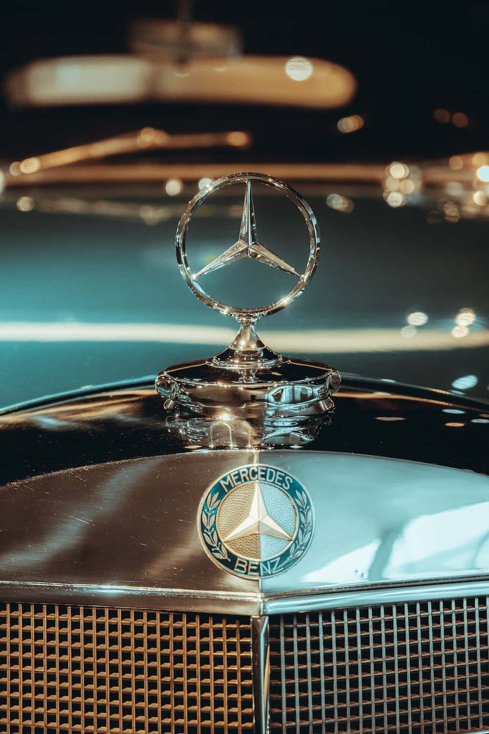 Mercedes 桌布 壁紙 — 夜間暖色散景燈光下閃爍的 vintage Mercedes-Benz 引擎蓋飾件特寫。