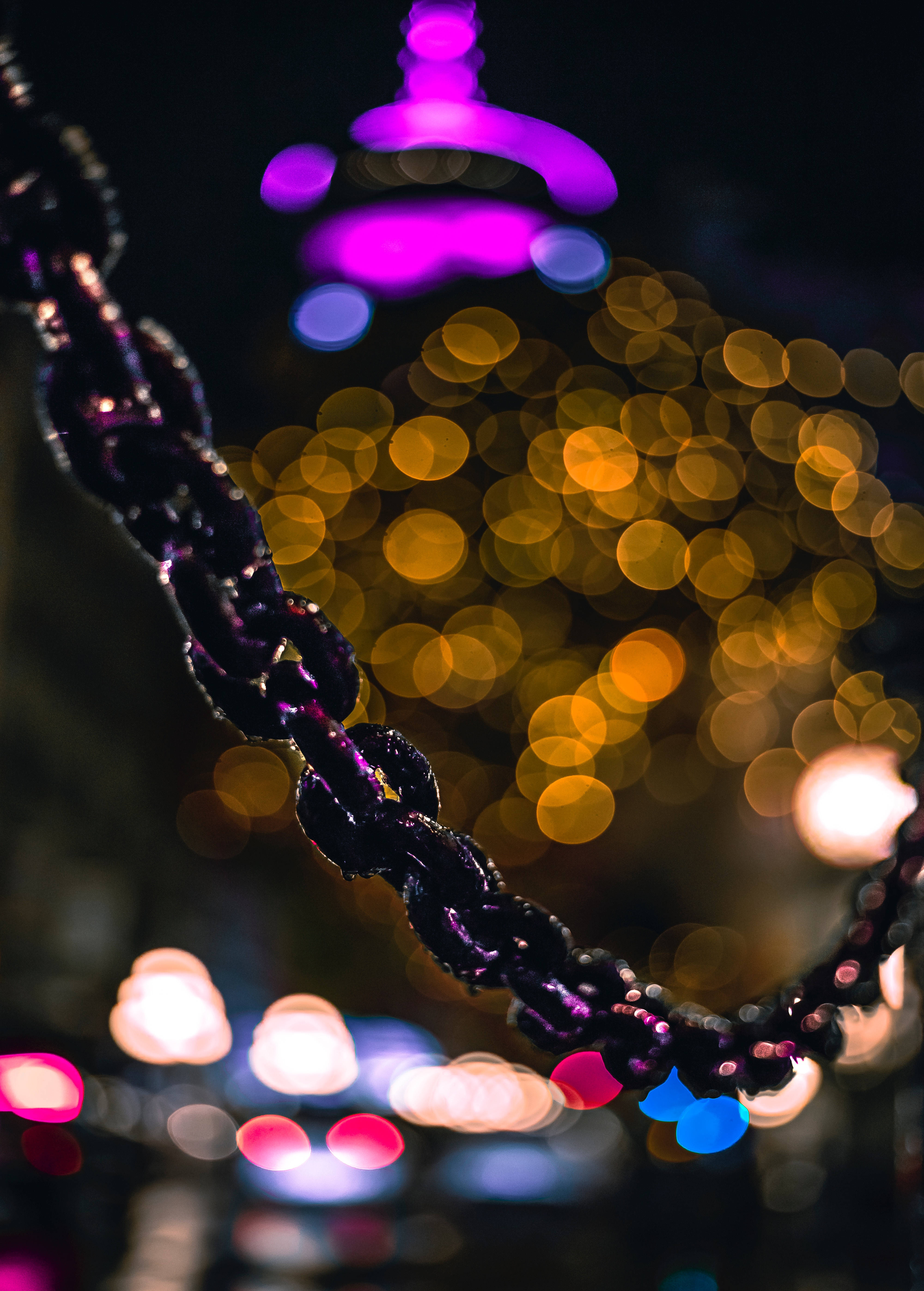 Bokeh 桌布 壁紙 — 夜間暖金色與冷紫色城市燈光散景背景下，帶有紫色高光的深色鏈條特寫。