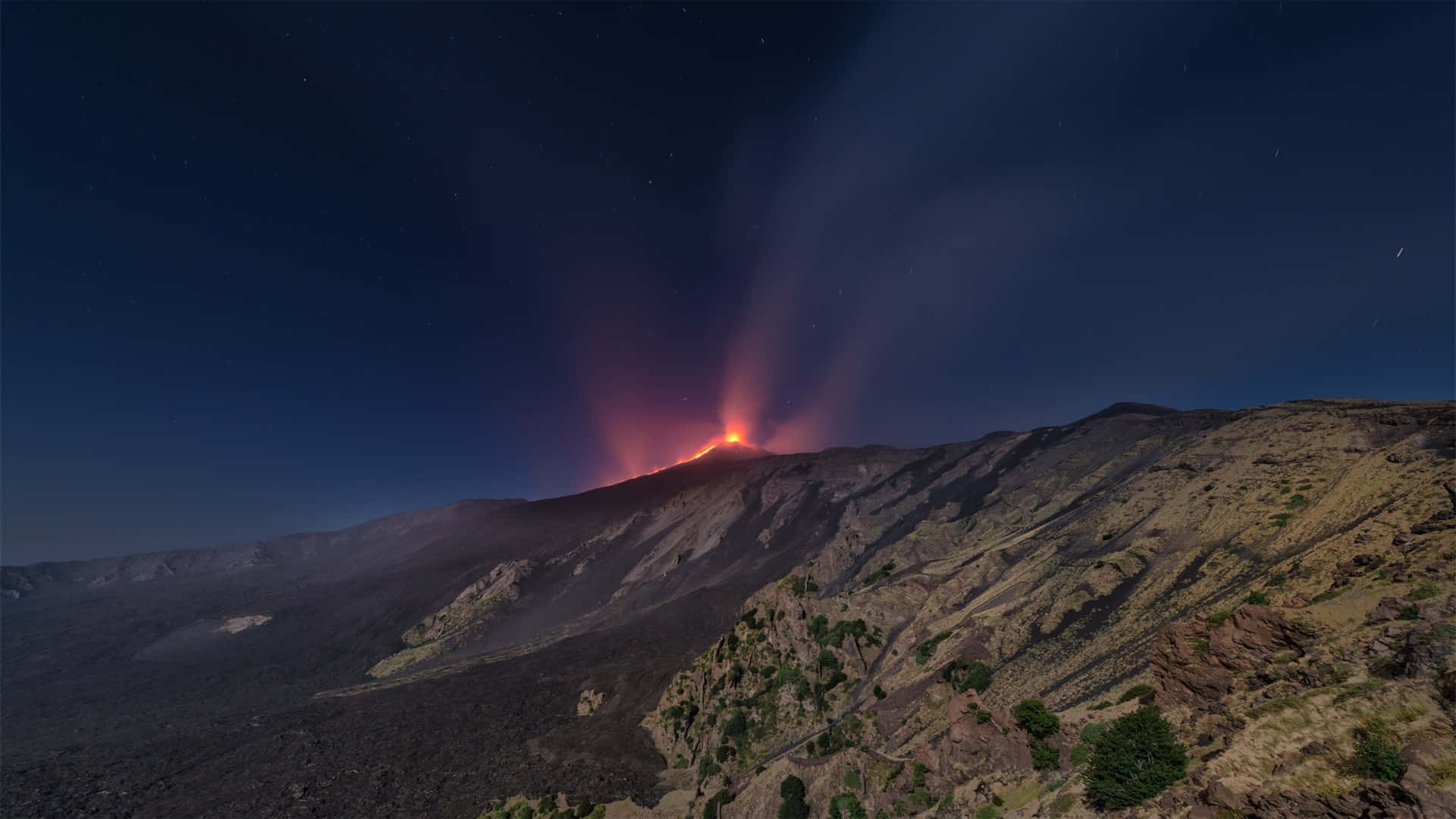 夜空 桌布 壁紙 — 夜間火山爆發照亮了崎嶇的山脈，在星空下伴隨著發光的熔岩和滾滾濃煙。