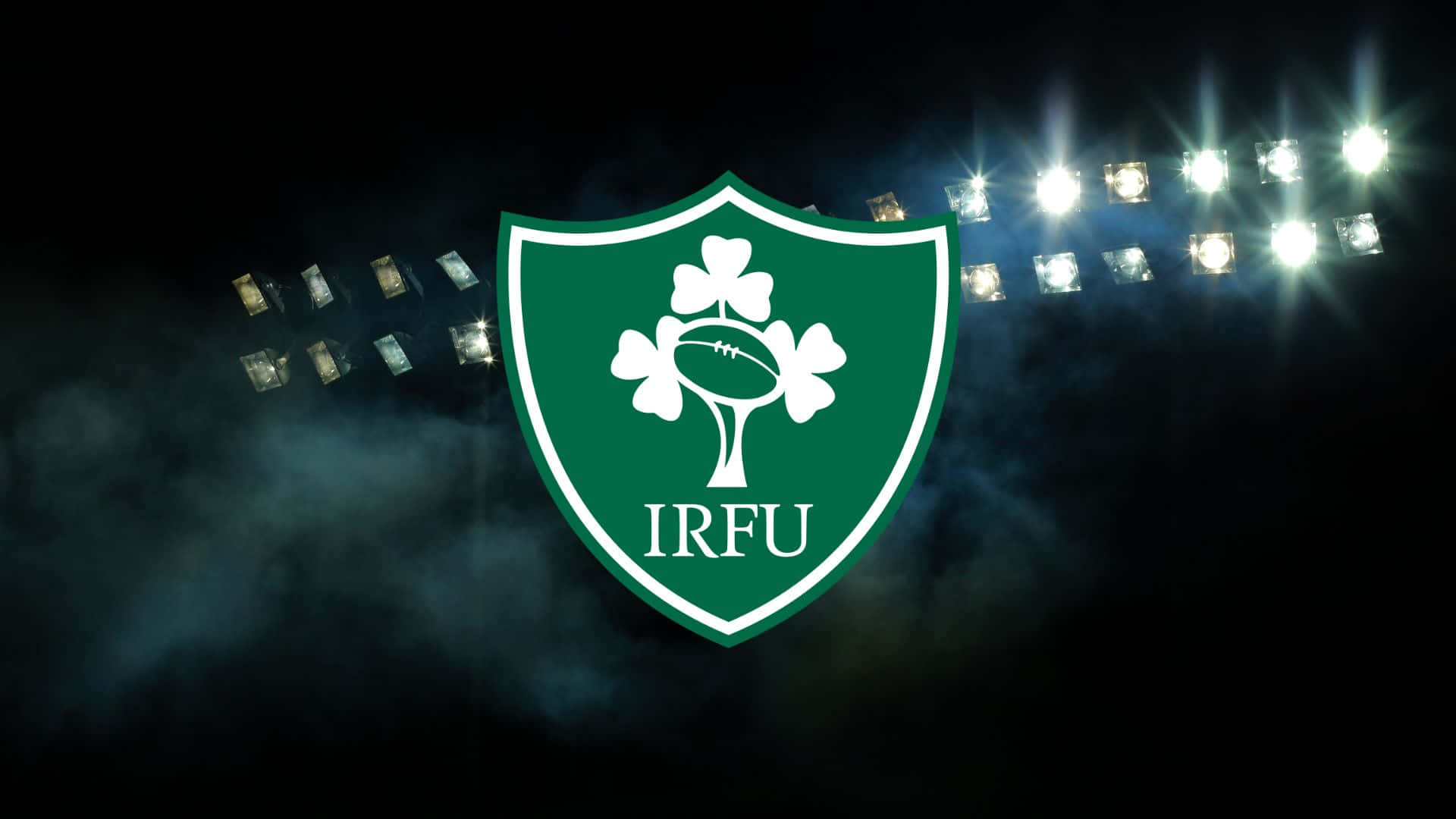 Ireland Rugby 桌布 壁紙 — 夜間球場燈光下，一個發光的綠色IRFU盾牌，中央有酢漿草和橄欖球。