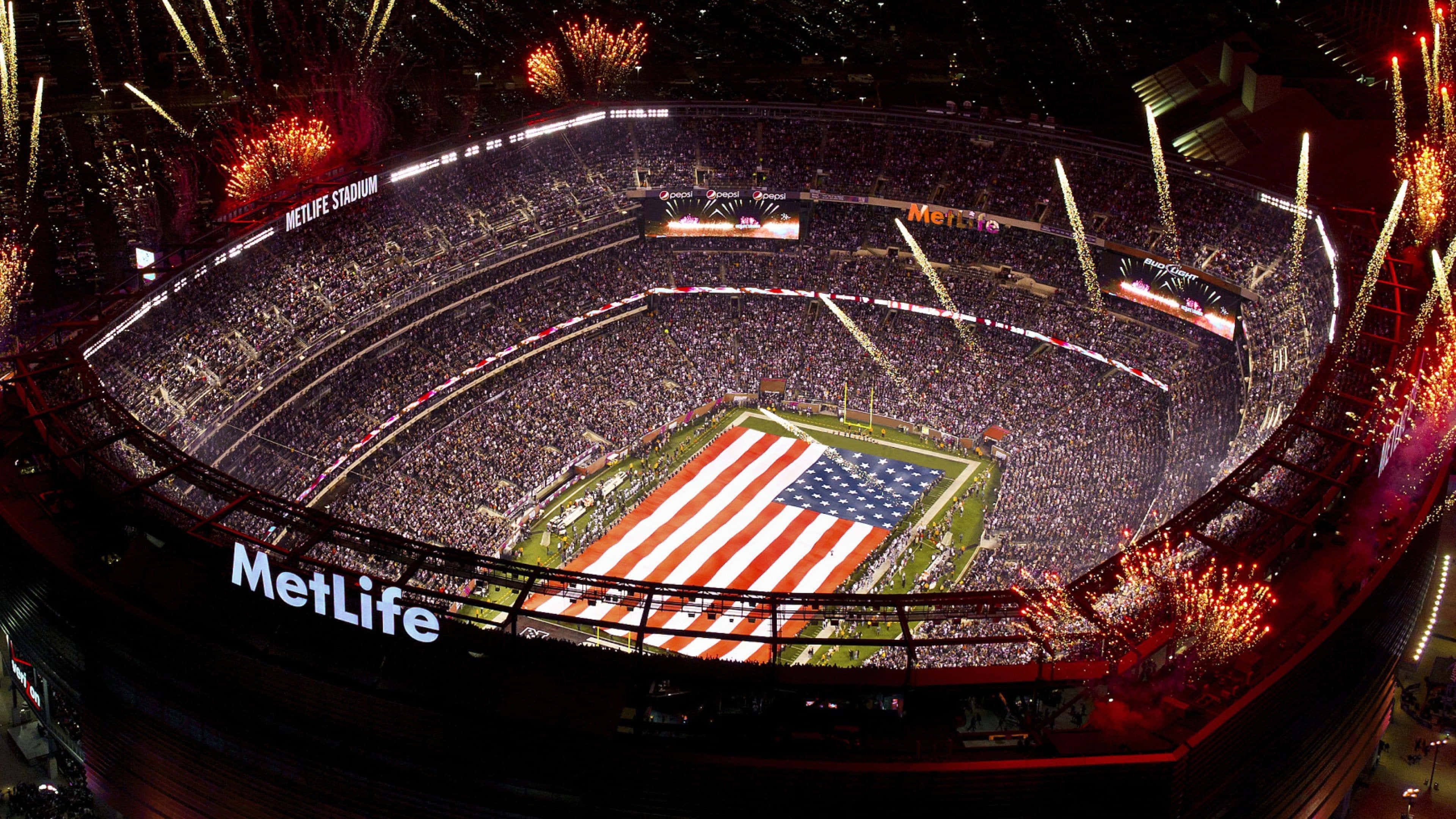 Super Bowl 桌布 壁紙 — 夜間美式足球場MetLife Stadium的鳥瞰圖，場內擠滿球迷，球場上空煙火綻放，顯示一面巨大的美國國旗。
