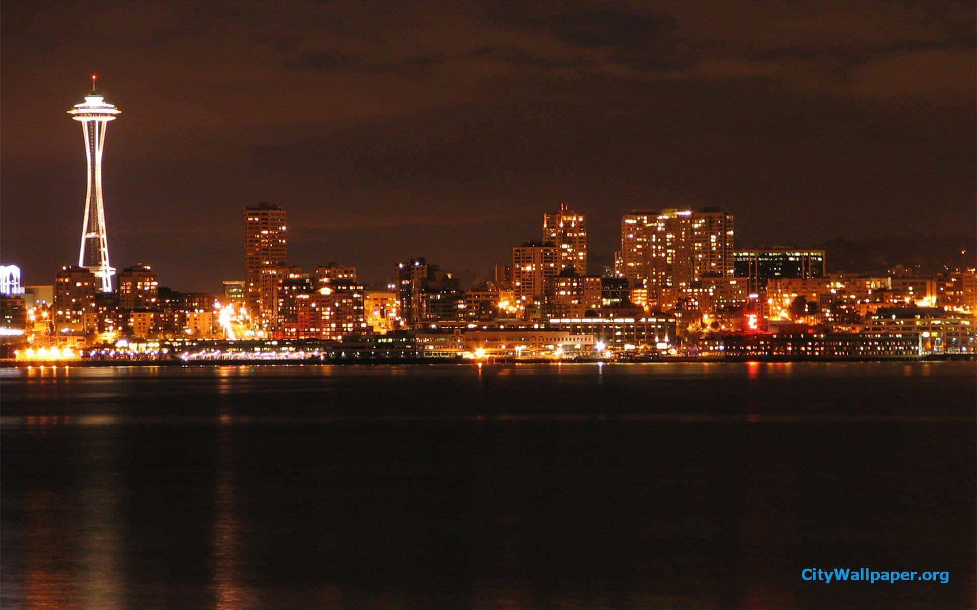 Seattle At Night 桌布 壁紙 — 夜間西雅圖天際線，太空針塔在水面上發光，倒映著城市燈光，天空烏雲密布。