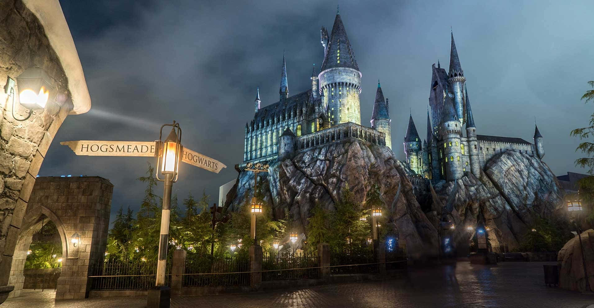 Hogsmeade 桌布 壁紙 — 夜間霍格華茲城堡亮起燈光，窗戶發光，並有一個指向活米村的路牌，背景是黑暗的天空。