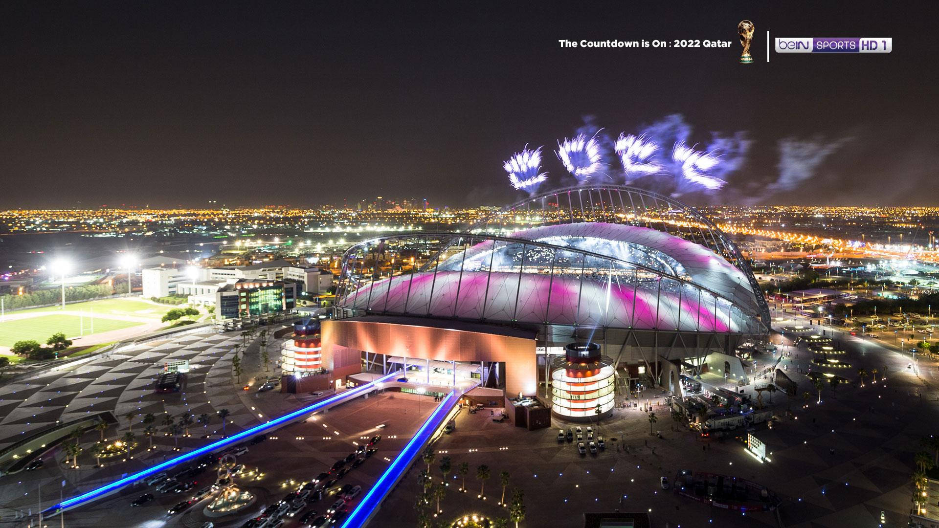 World 桌布 壁紙 — 夜間鳥瞰發光體育場，上空有「QATAR」煙火，周圍是城市燈光和停車場。