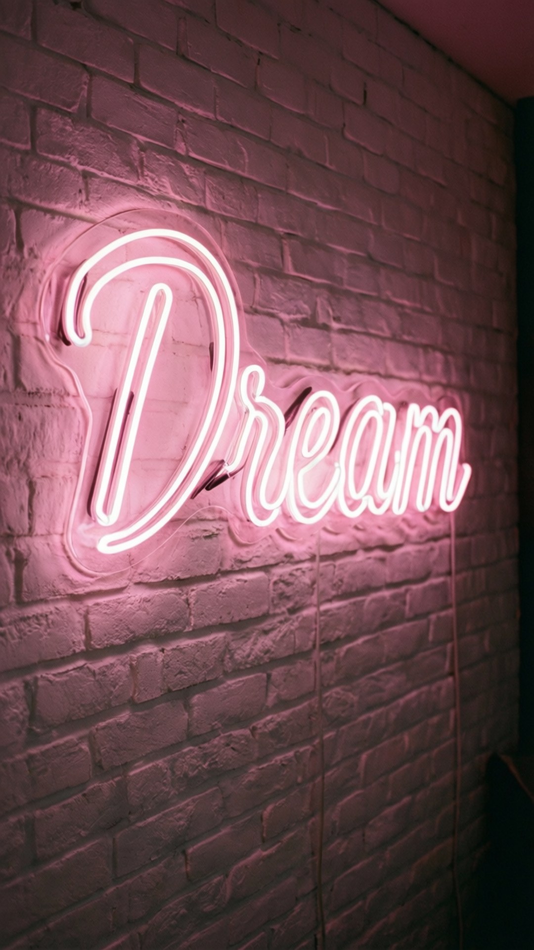 夢境霓虹燈 壁紙 — 一個發光的粉紅色霓虹燈牌，拼出「Dream」字樣，在昏暗的房間中將柔和的光線投射在有紋理的磚牆上。