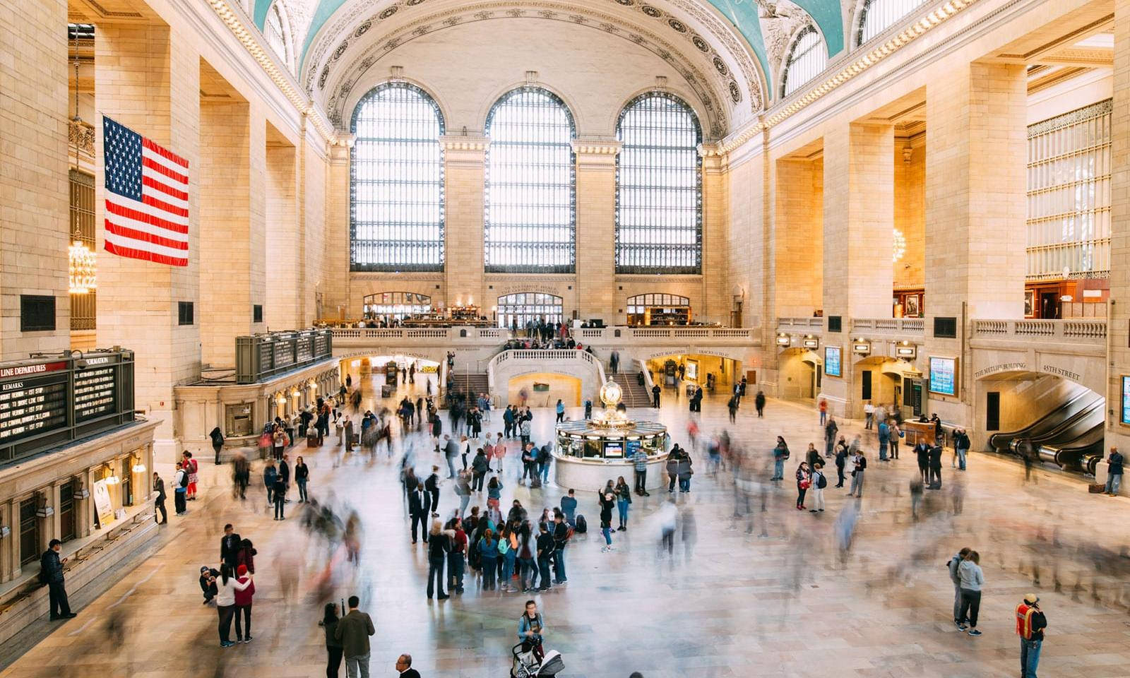 New York 桌布 壁紙 — 大中央車站高聳的石砌大廳在拱形窗戶和巨型美國國旗下，擠滿了模糊的旅客。