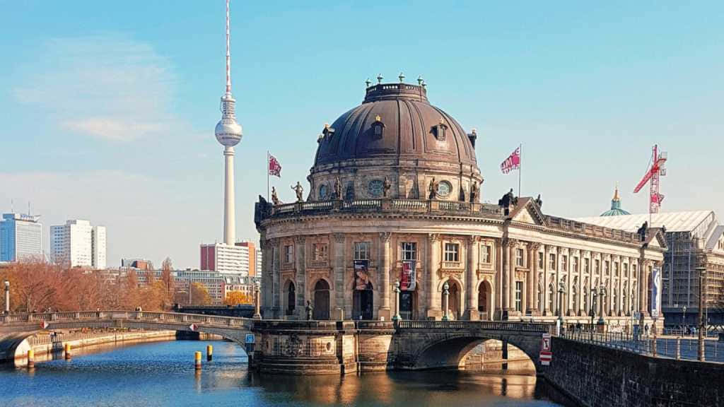 Berlin 桌布 壁紙 — 大型圓頂建築帶旗幟矗立在河岸上，在清澈的藍天下，背景可見柏林電視塔。