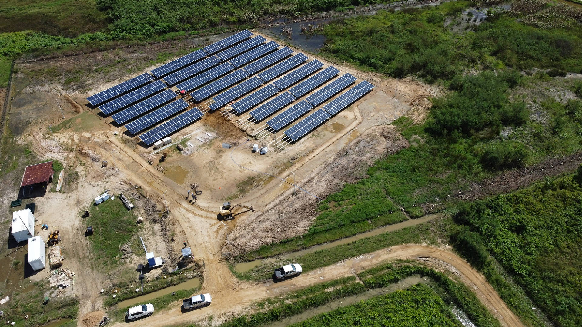 電力 桌布 壁紙 — 大型太陽能發電廠正在建設中的鳥瞰圖，周圍環繞著茂密的綠色植被和帶有車輛的泥路。