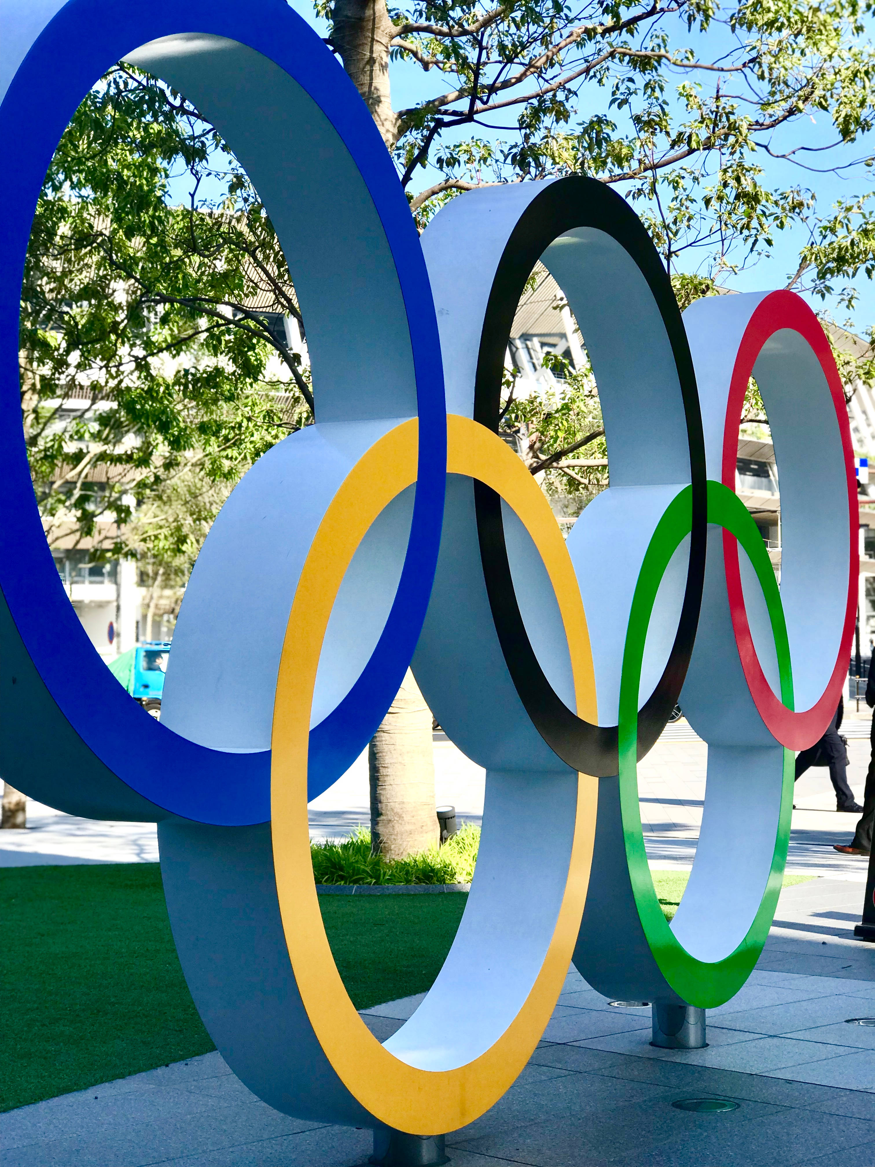 Olympics 桌布 壁紙 — 大型奧運五環雕塑色彩鮮豔，矗立在戶外，藍天白雲下，背景有綠樹和城市建築。