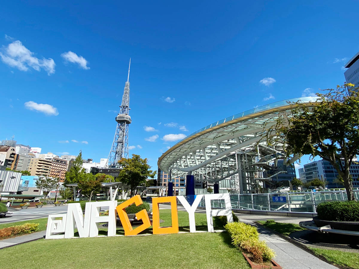 Nagoya 桌布 壁紙 — 大型彩色NAGOYA字母放置在草地上，背景是明亮的藍天、散落的雲朵和名古屋電視塔。