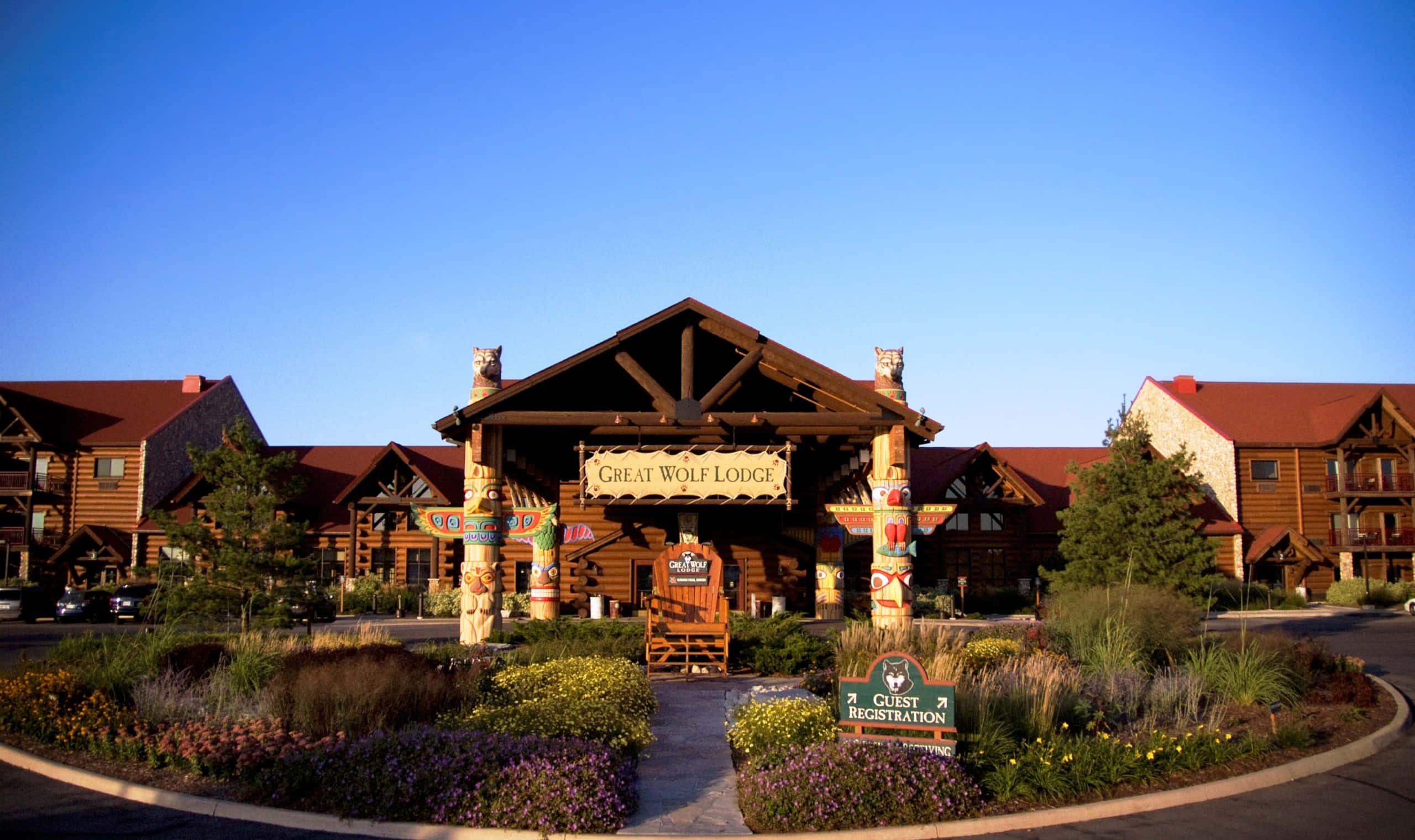 great wolf lodge 桌布 壁紙 — 大型木製小屋入口，雕刻柱子和色彩繽紛的花圃，在清澈藍天下。