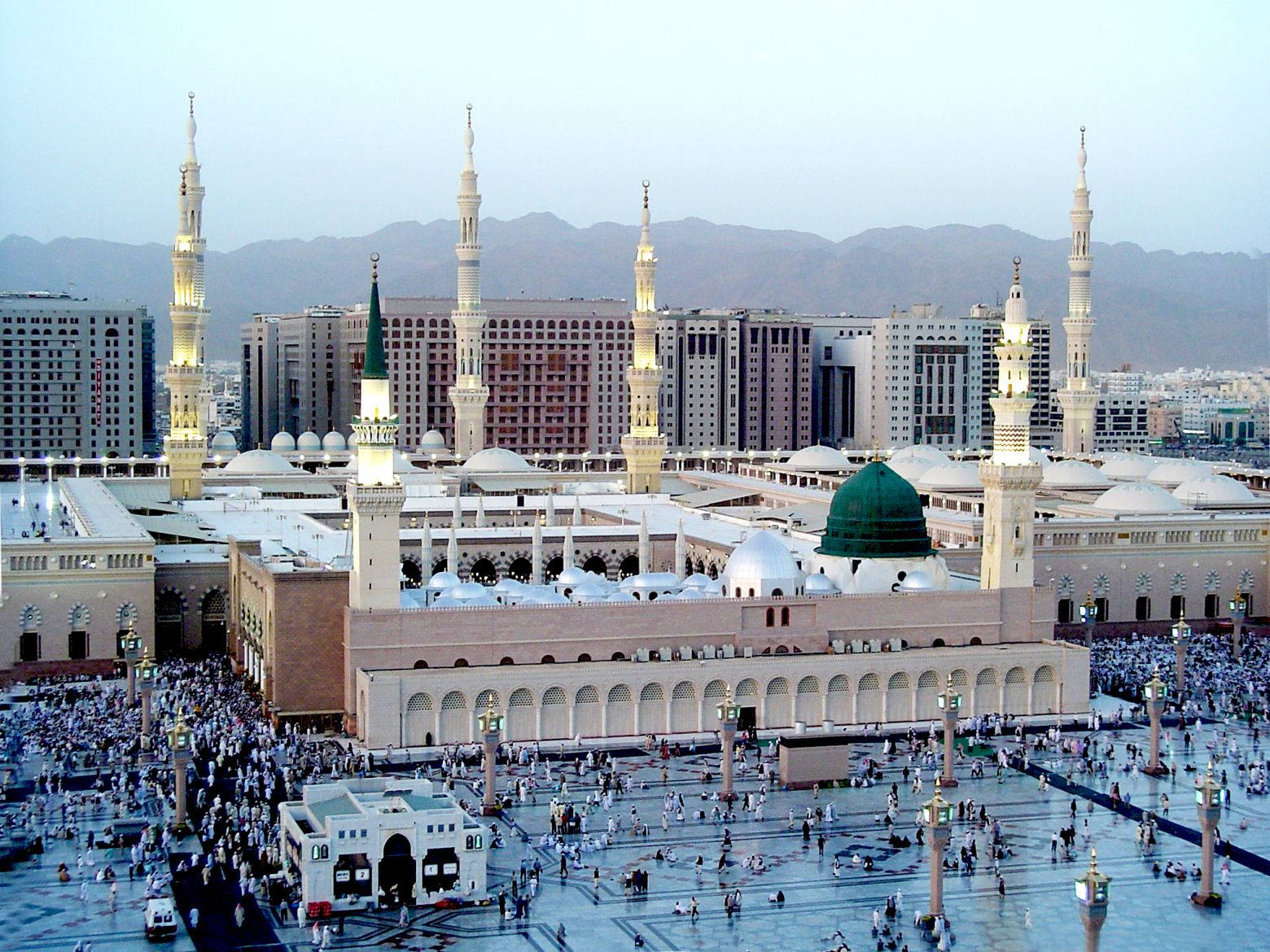 Madina Sharif 桌布 壁紙 — 大型清真寺，擁有綠色圓頂和高聳的宣禮塔，在黃昏時分被廣闊庭院中的人群包圍。