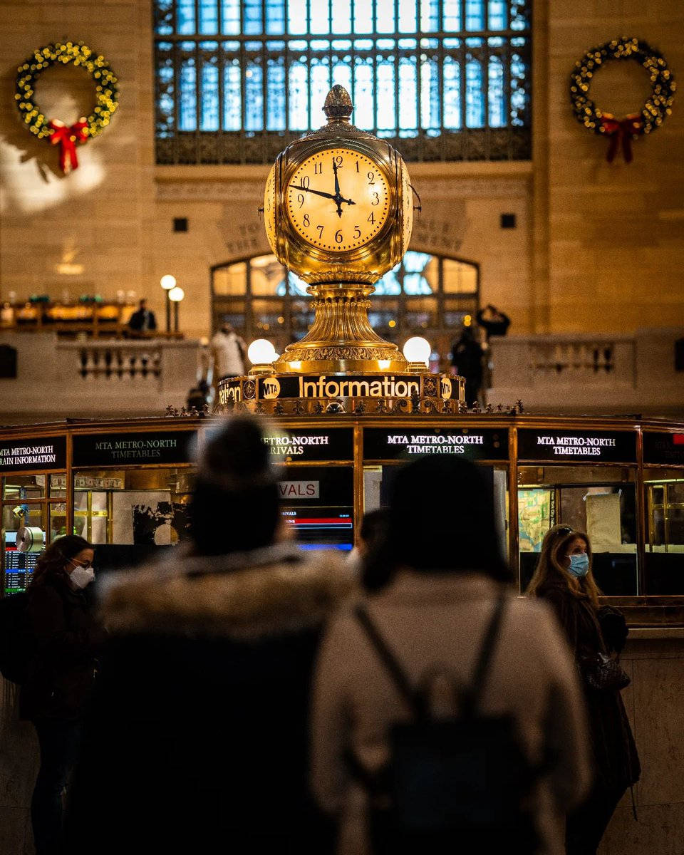 New York 桌布 壁紙 — 大型火車站時鐘在溫暖燈光下閃耀，戴著口罩的旅客穿過裝飾著節日花環的歷史大廳。