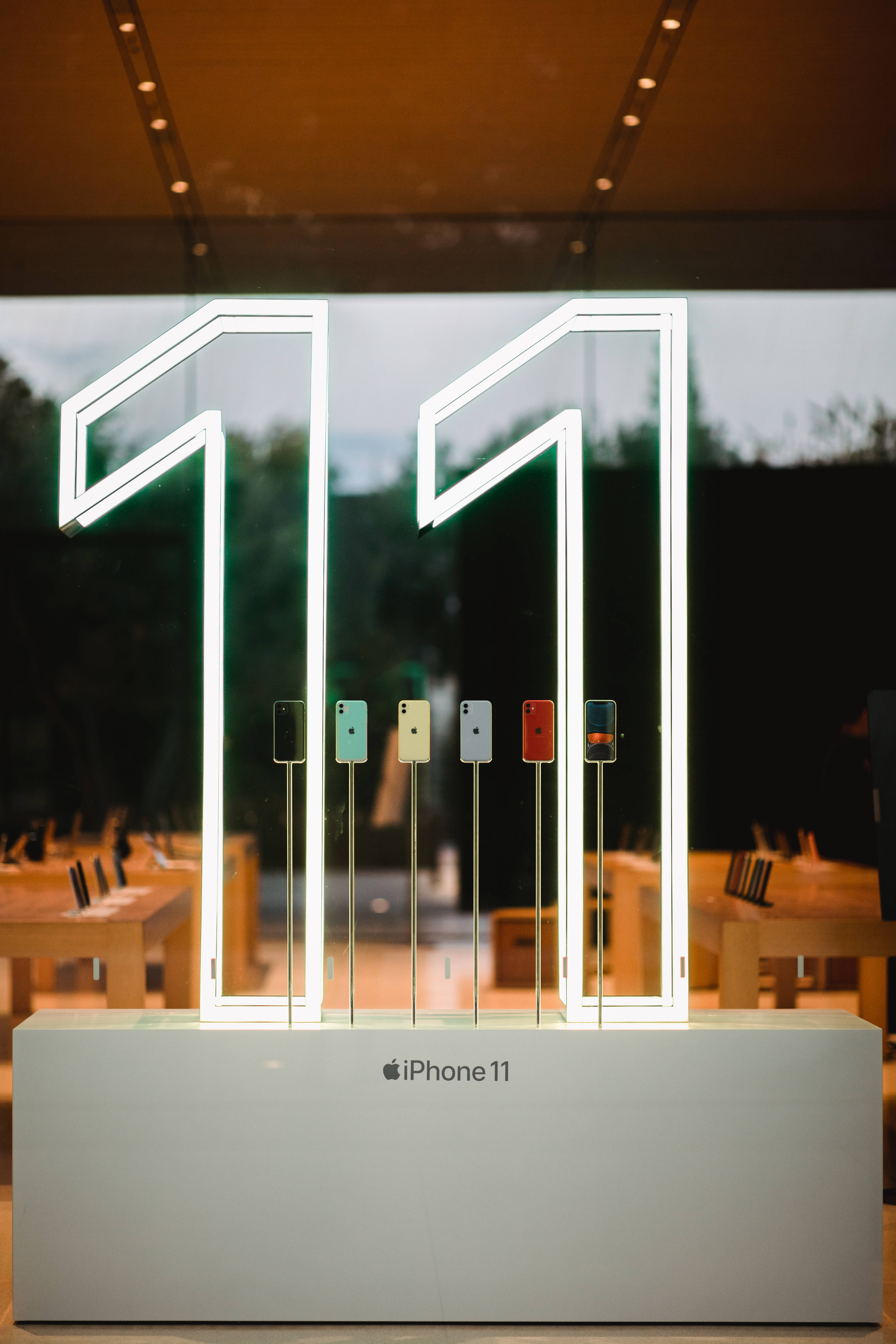 iPhone 11 桌布 壁紙 — 大型發光霓虹數字11框住色彩繽紛的iPhone 11型號，展示在現代Apple Store內的時尚支架上。