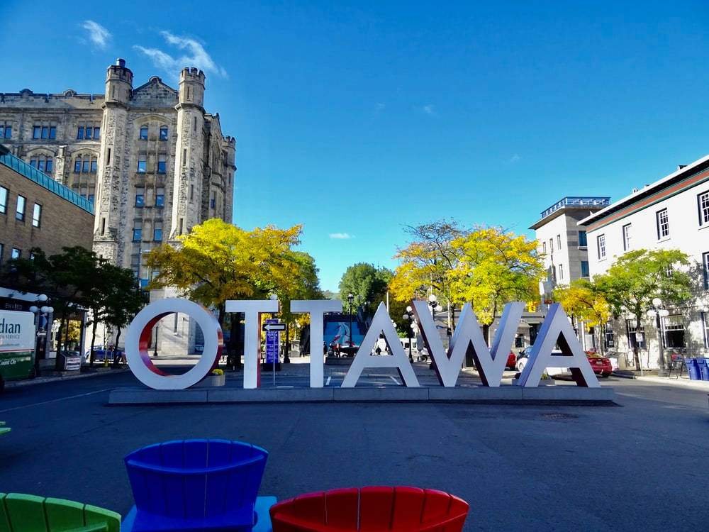 Market 桌布 壁紙 — 大型白色「OTTAWA」字母矗立在陽光明媚的城市廣場上，周圍有秋樹和歷史建築，背景是清澈的藍天。