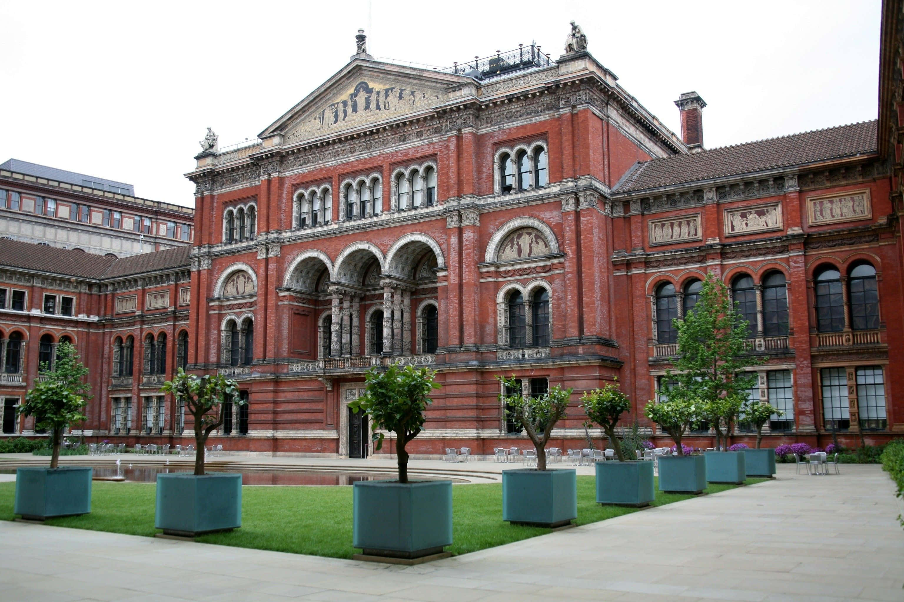 Museums 桌布 壁紙 — 大型紅磚維多利亞式博物館，擁有拱形窗戶和雕像，矗立在修剪整齊的草坪和盆栽樹木之後。