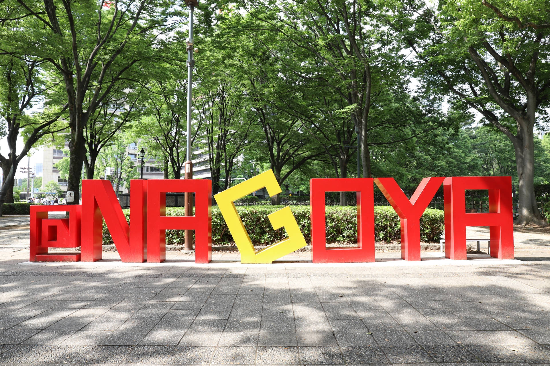Nagoya 桌布 壁紙 — 大型紅色和黃色的 '@NAGOYA' 字母矗立在陽光明媚的公園中，周圍環繞著茂密的綠樹和鋪砌的小徑。