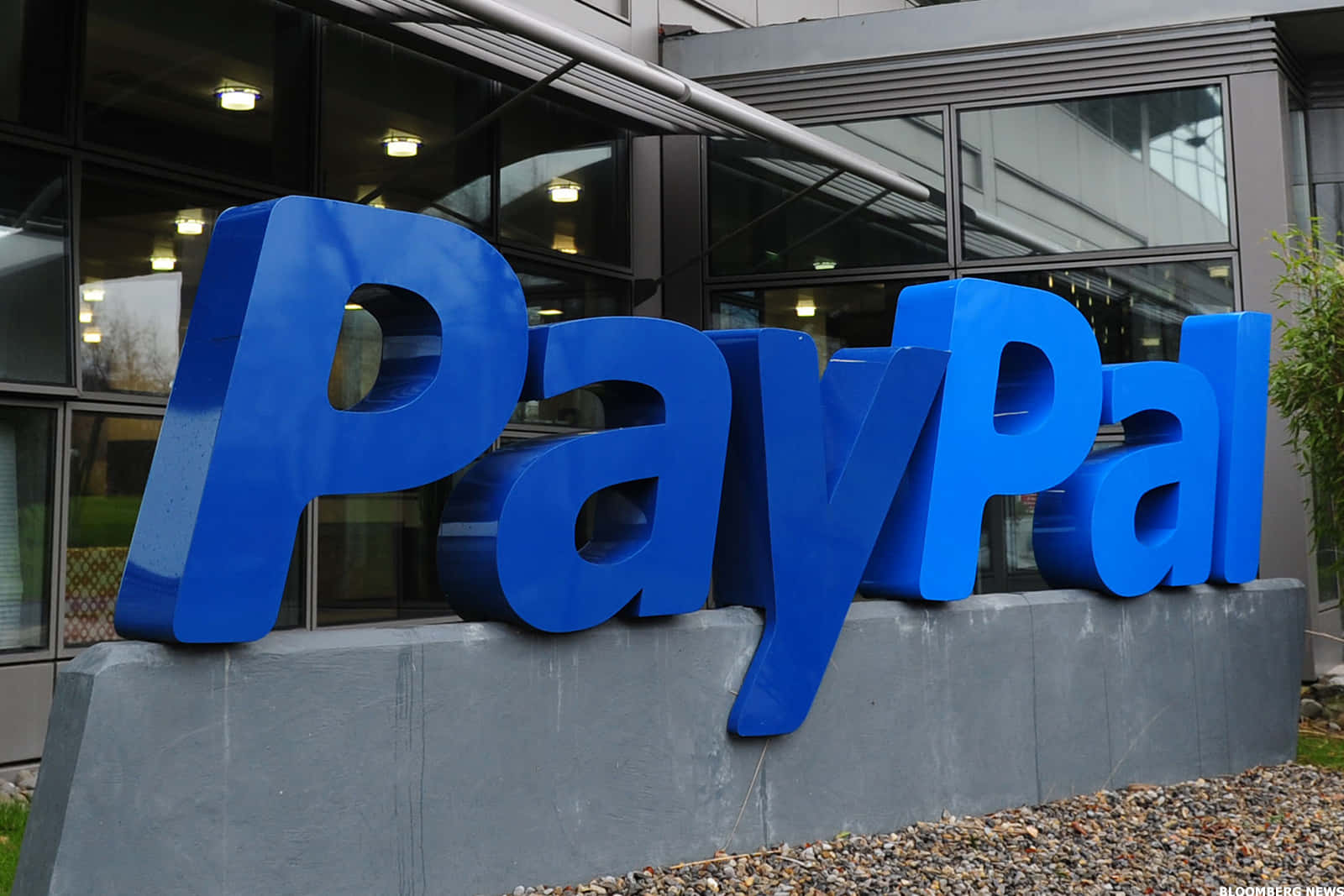PayPal 桌布 壁紙 — 大型藍色PayPal標誌字母放置在現代玻璃建築外的灰色底座上，地面鋪有礫石。