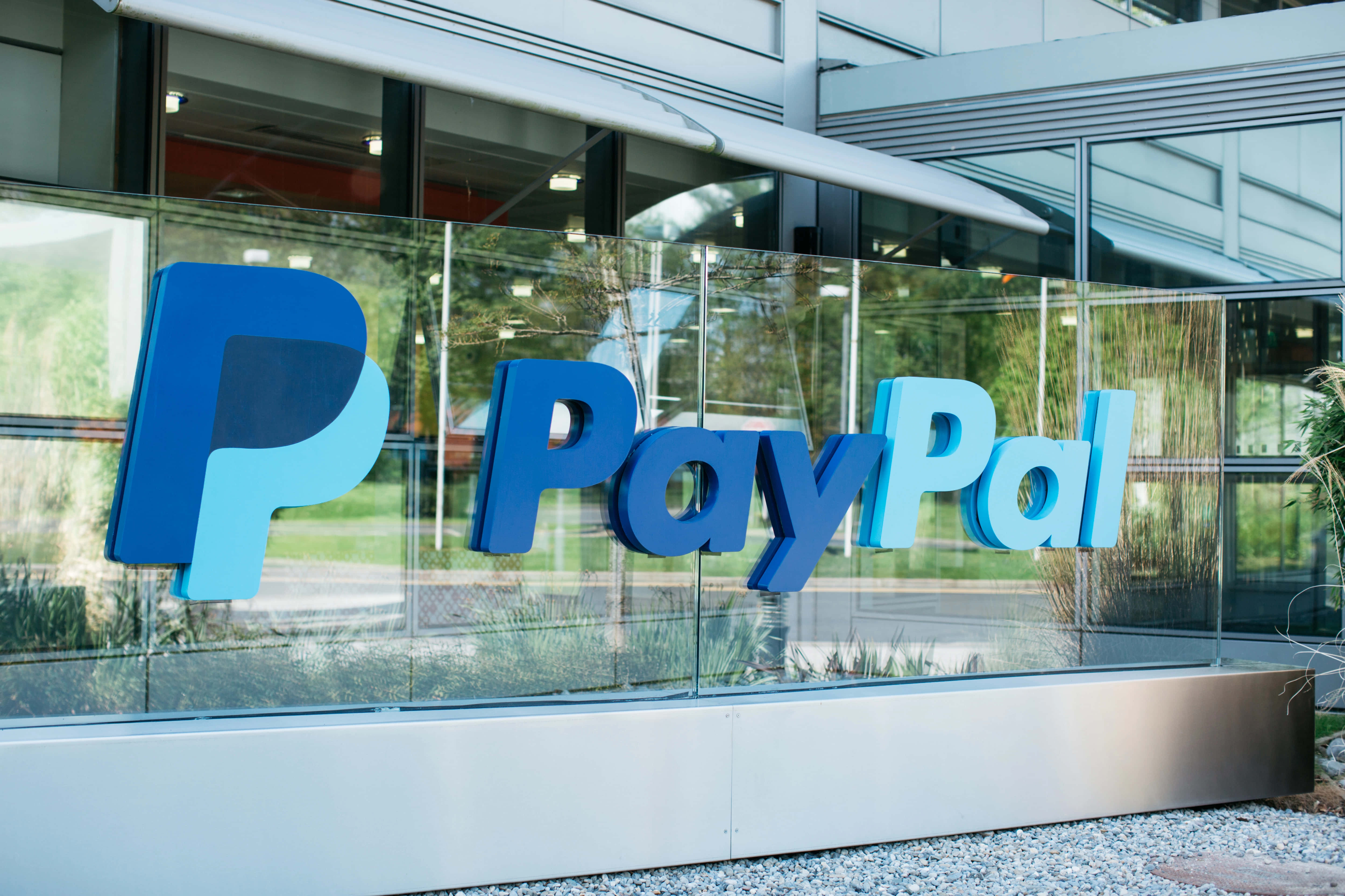 PayPal 桌布 壁紙 — 大型藍色PayPal標誌安裝在現代辦公大樓的玻璃外牆上，地面鋪有碎石並映照綠意。