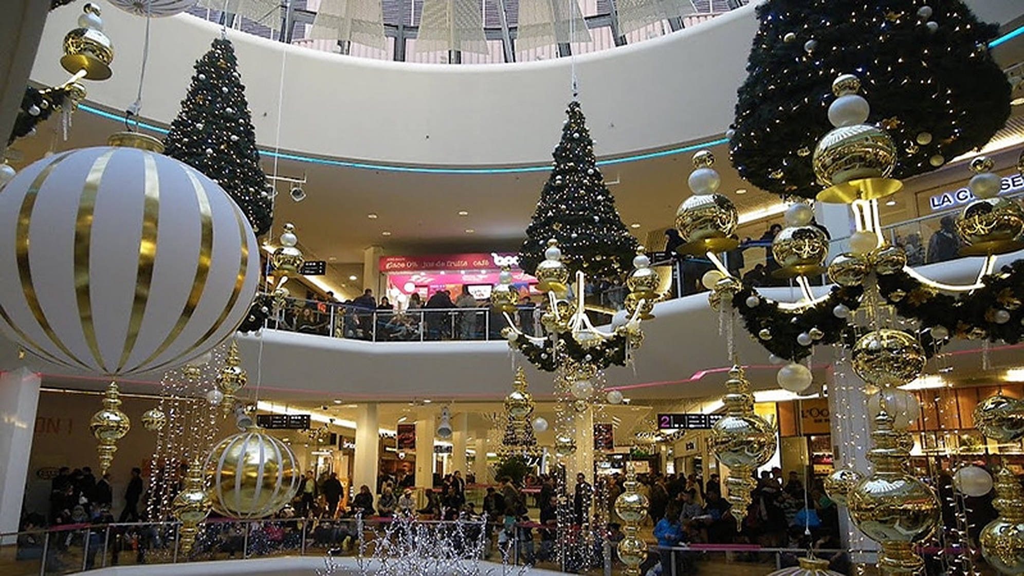 Mall 桌布 壁紙 — 大型購物中心中庭，裝飾著高聳的黑色聖誕樹和懸掛在弧形天花板上的超大型金色和白色飾品。