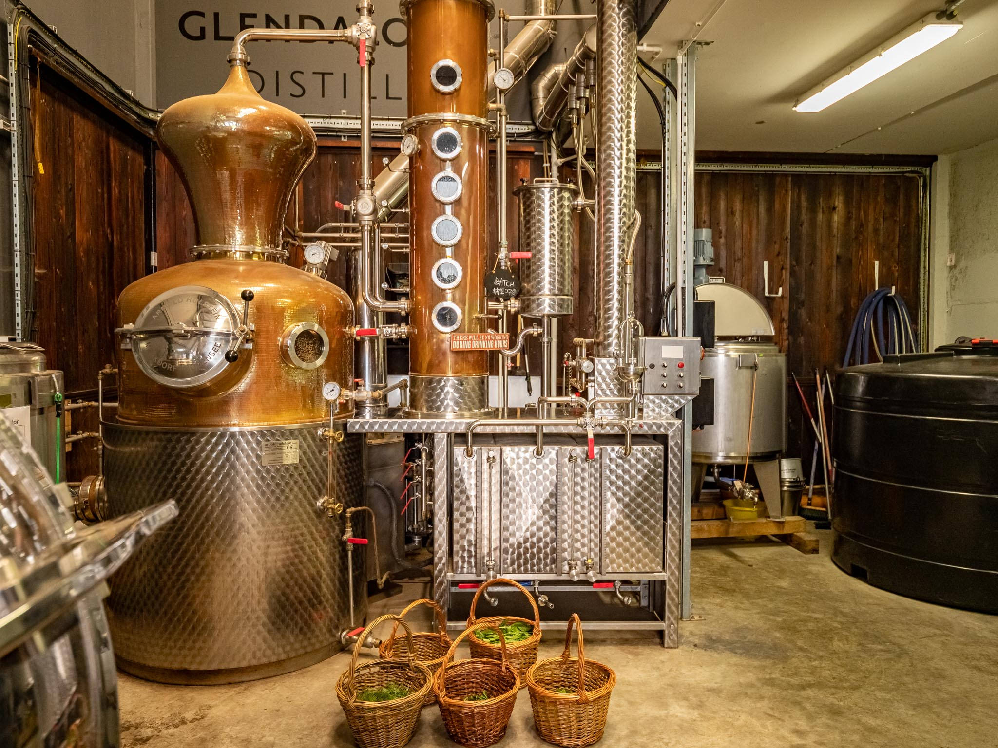 Whiskey 桌布 壁紙 — 大型銅和不鏽鋼蒸餾設備主導著一個質樸的蒸餾室，前景有編織的綠色香草籃。