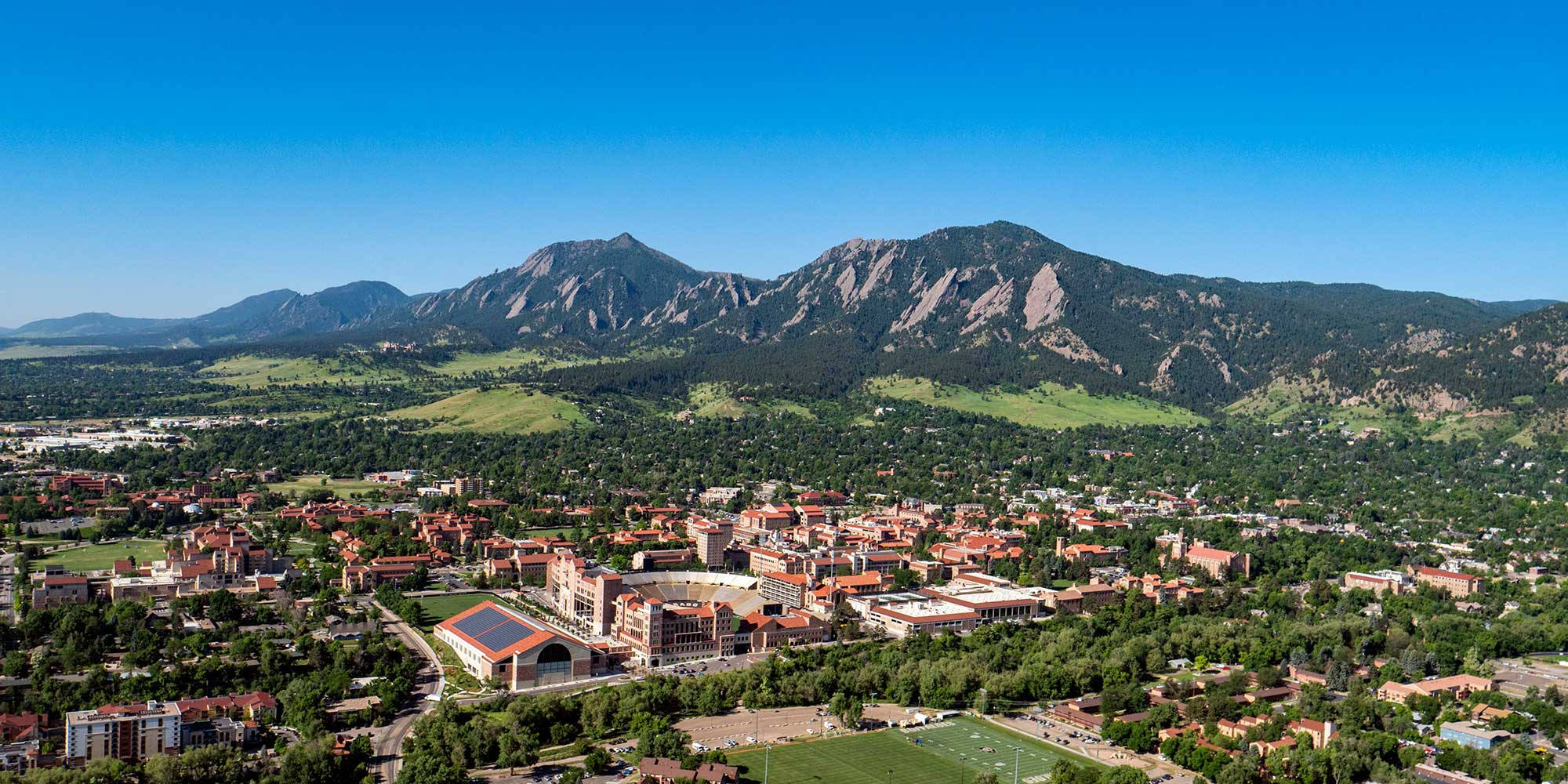 University Of Colorado 桌布 壁紙 — 大學城的鳥瞰圖，坐落在翠綠的山谷中，突出的岩石山脈在晴朗的藍天下聳立。