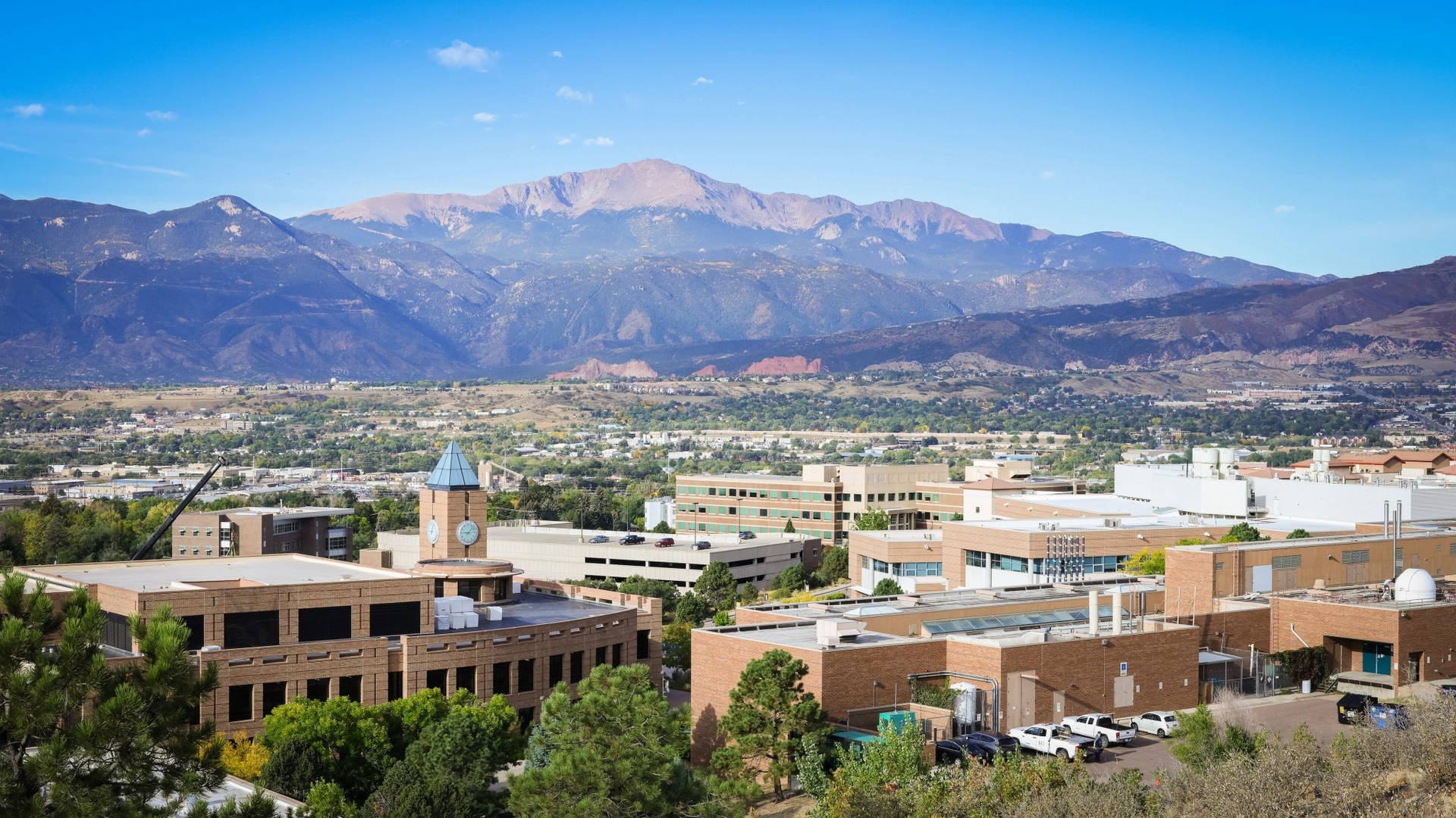  壁紙 — 大學校園的全景，磚砌建築和鐘樓坐落在綠樹叢中，背景是晴朗藍天下雄偉的雪山。