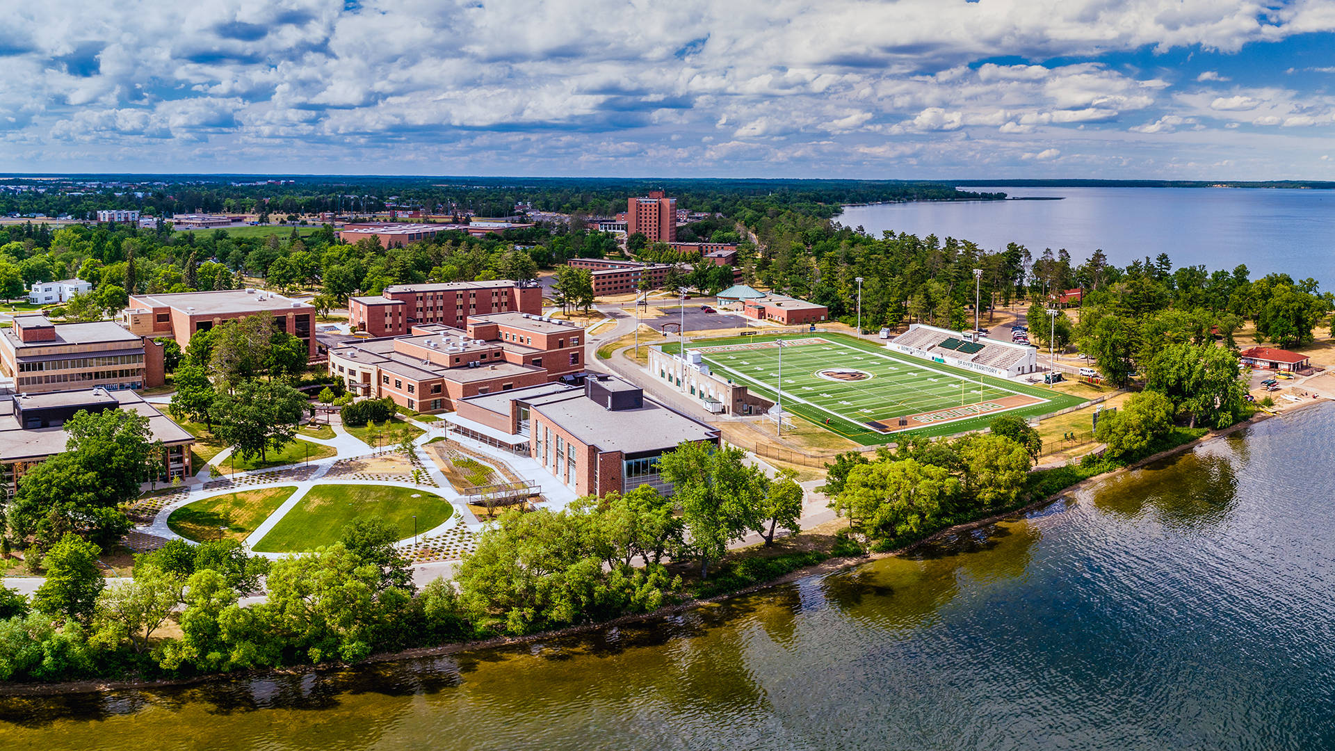 Minnesota 桌布 壁紙 — 大學校園的鳥瞰圖，有磚砌建築、綠色草坪和一個位於大湖旁邊的足球場，天空部分多雲。