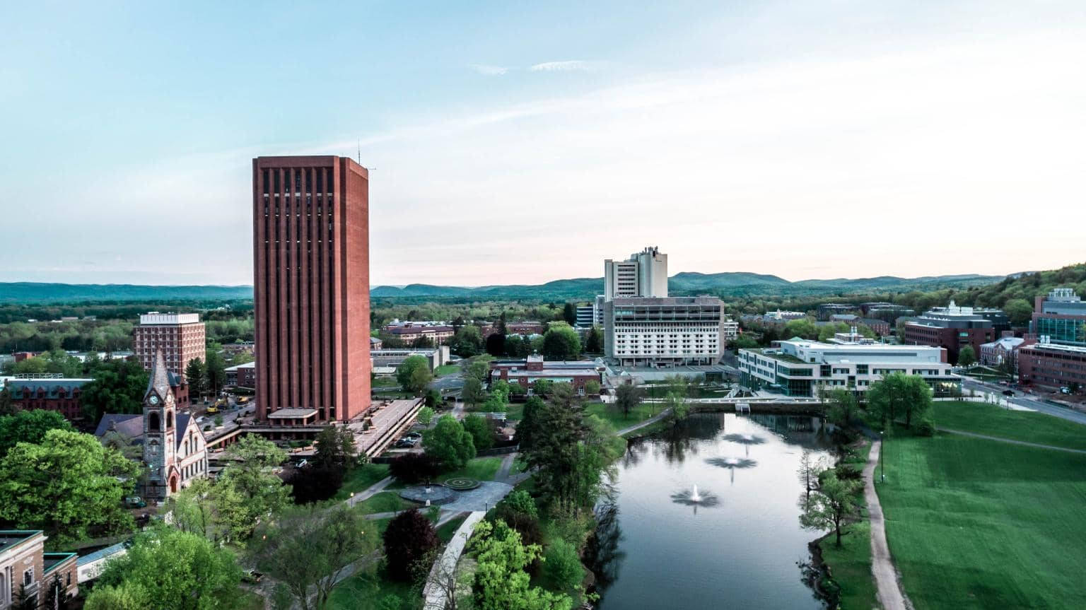 Building 桌布 壁紙 — 大學校園的鳥瞰圖，有紅磚塔、綠色草坪、平靜的河流和噴泉，天空柔和。