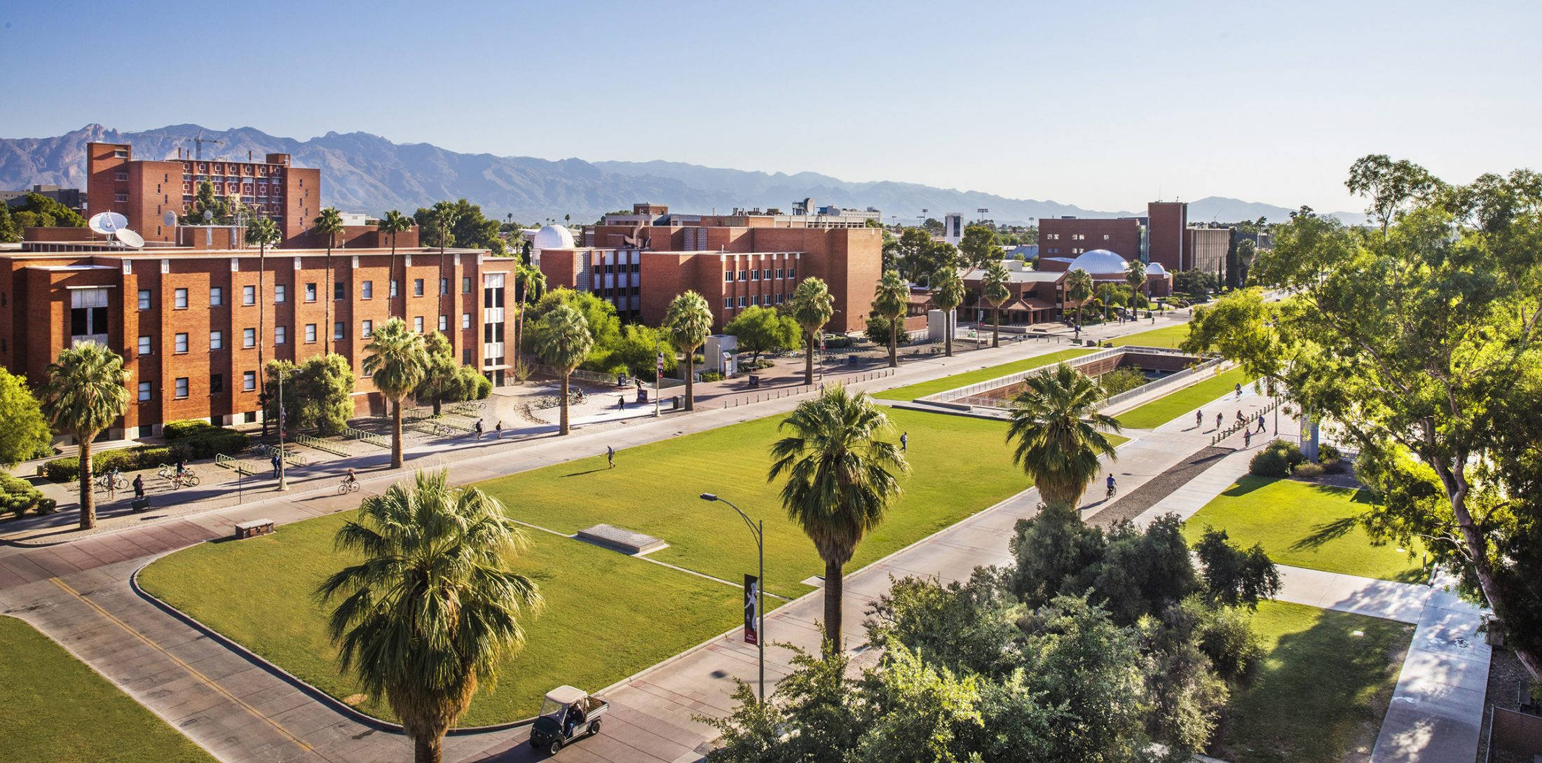 Arizona 桌布 壁紙 — 大學校園的鳥瞰圖，有紅磚建築、棕櫚樹和山脈背景，天空清澈湛藍。