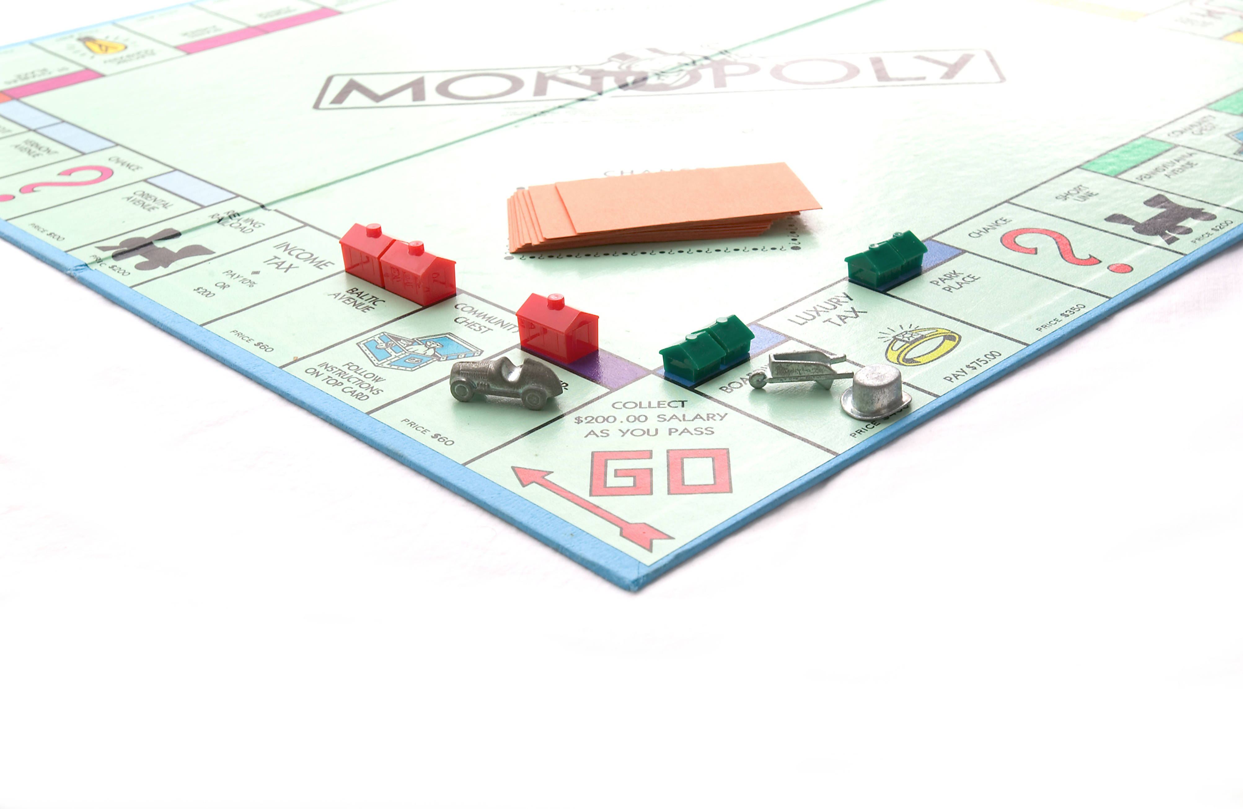 Monopoly 桌布 壁紙 — 大富翁遊戲板的特寫，上面有紅色和綠色的塑膠棋子，以及一堆橙色卡牌，放在白色表面上。