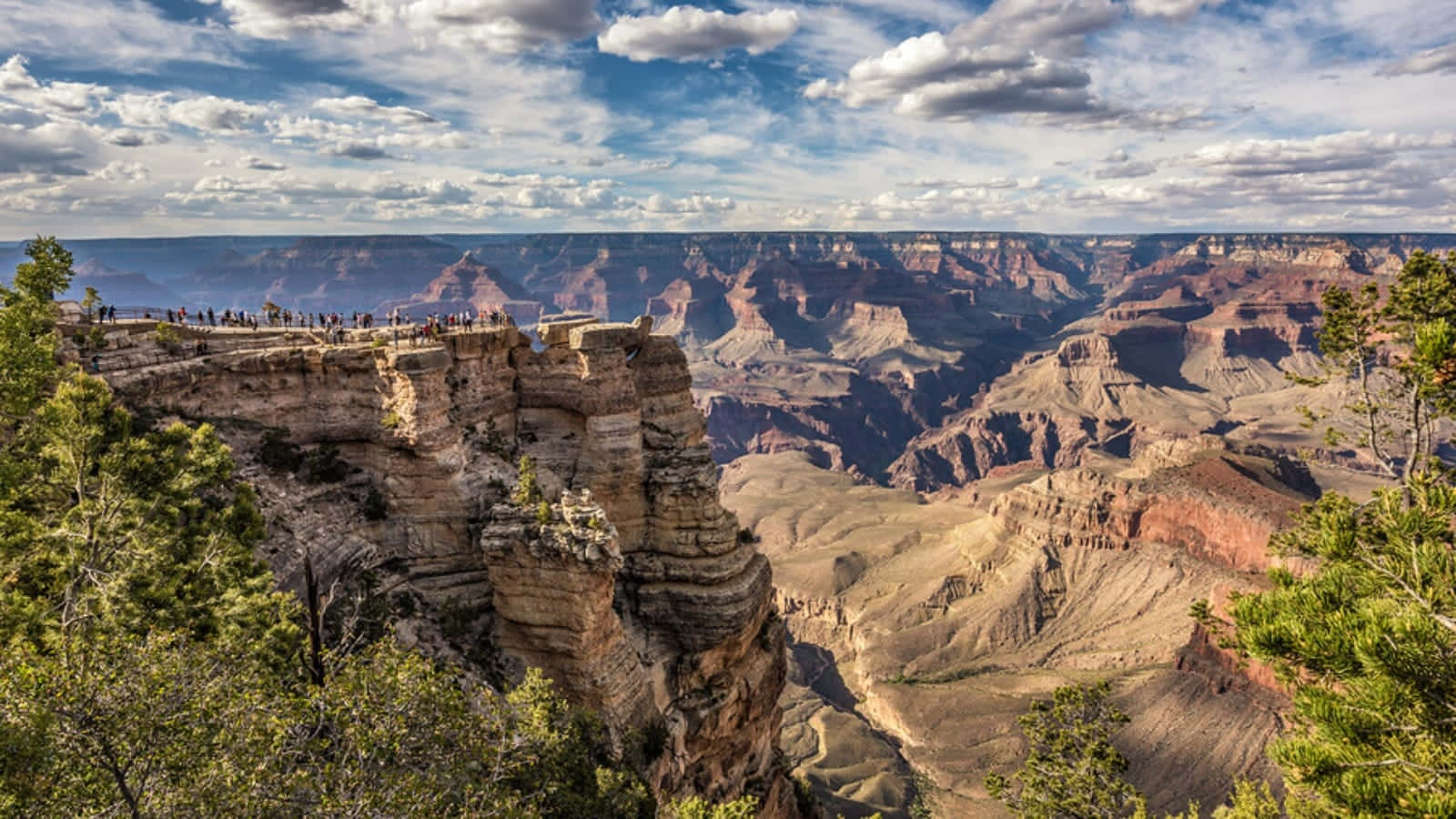 grand canyon 桌布 壁紙 — 大峽谷的壯麗景色，層層疊疊的紅色岩石形成在部分多雲的藍天下，邊緣有遊客。