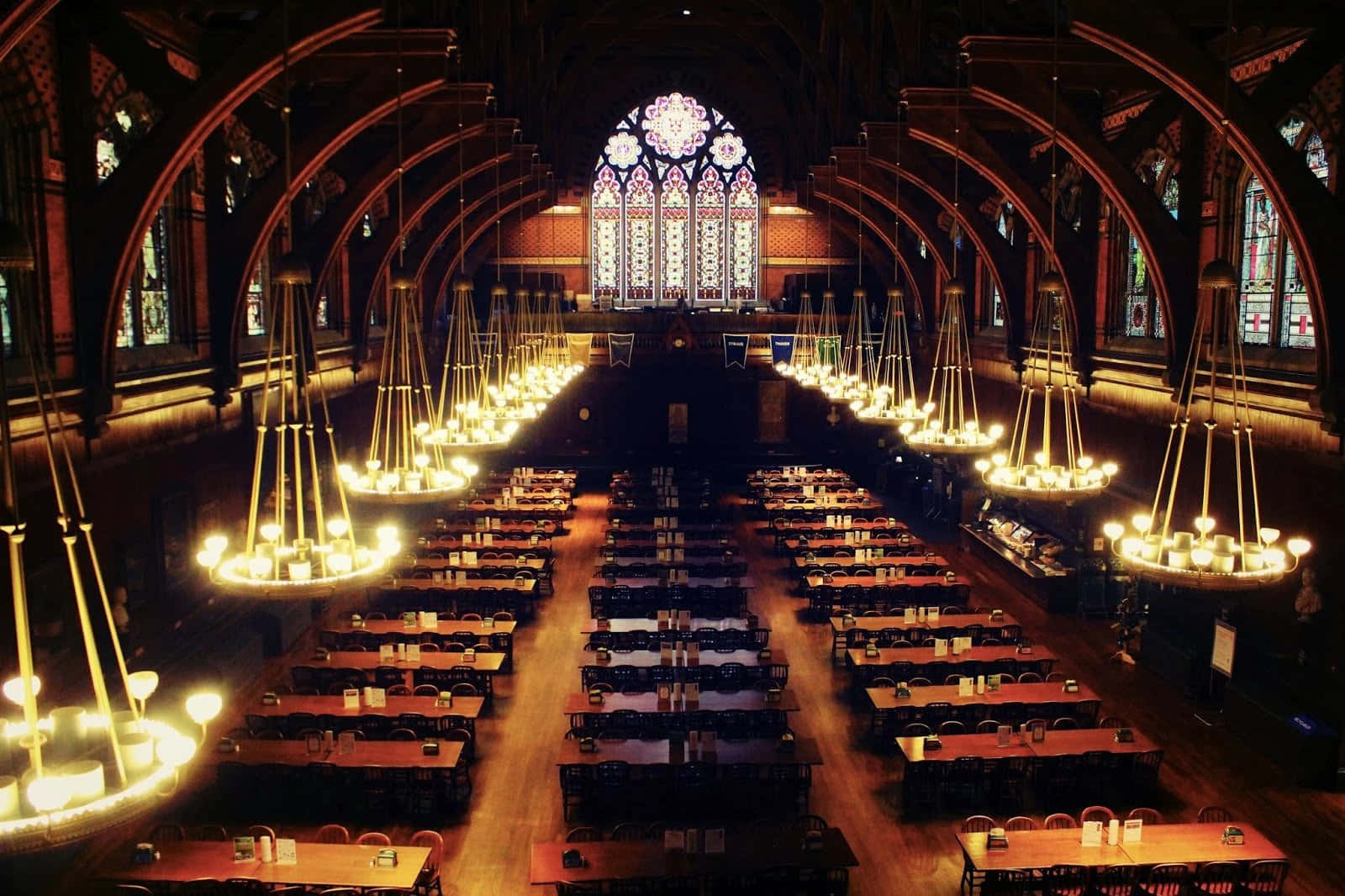 The Hogwarts Great Hall 桌布 壁紙 — 大教堂風格的大型餐廳，配有拱形彩色玻璃窗和發光的吊燈，照亮長木桌。
