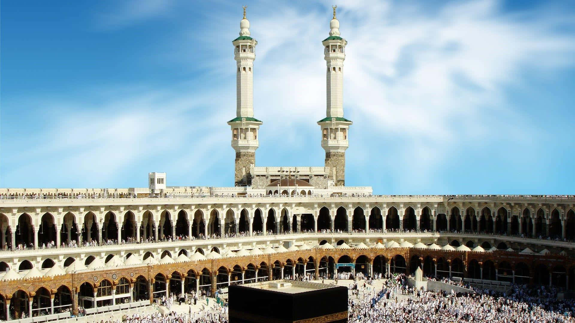 Kaaba 桌布 壁紙 — 大清真寺，有兩座高聳的宣禮塔和黑色天房，在明亮的藍天下被人群圍繞。