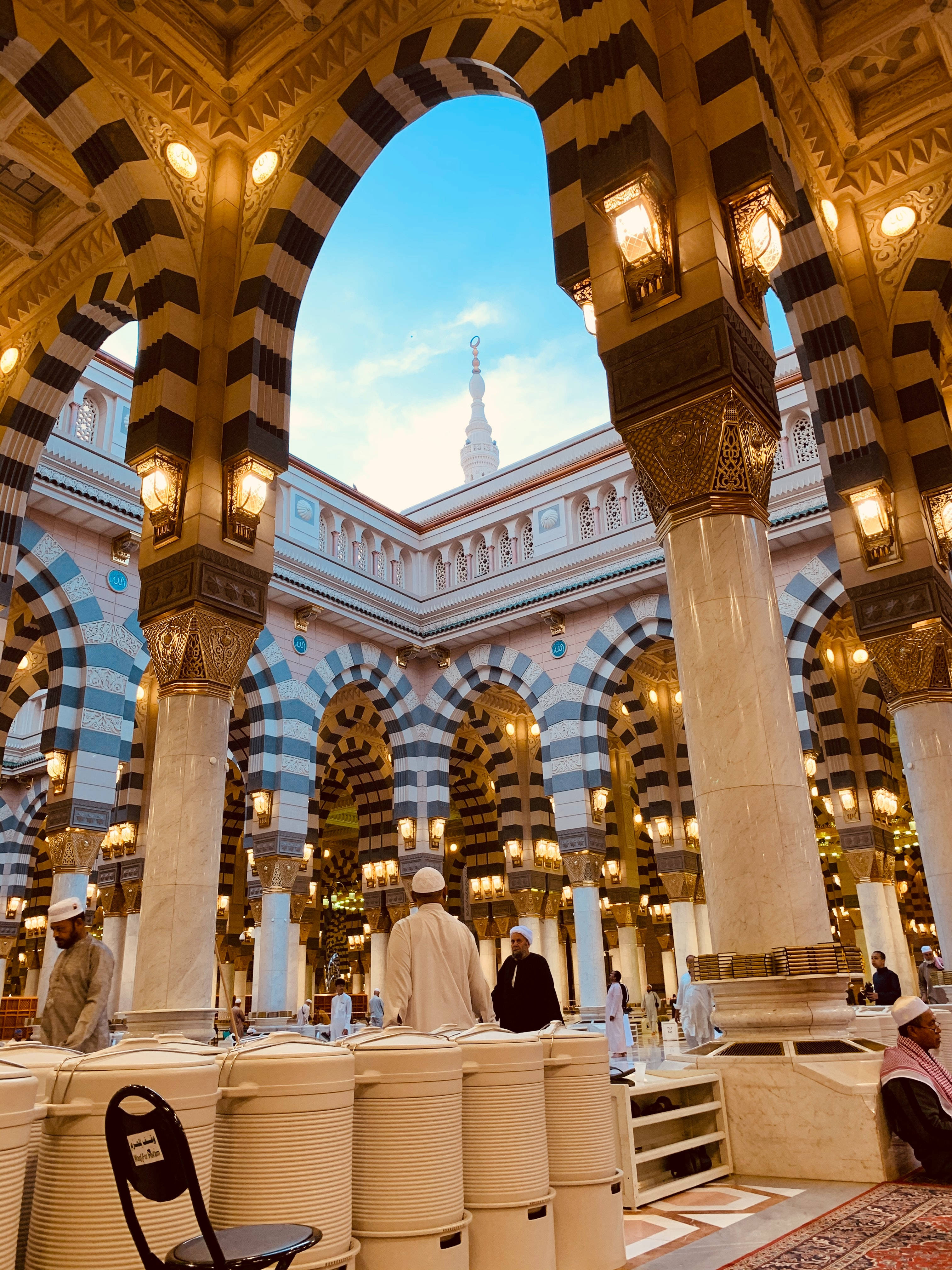 Madina 桌布 壁紙 — 大清真寺內部，條紋拱門在溫暖燈光下閃耀，信徒在晴朗藍天下祈禱。