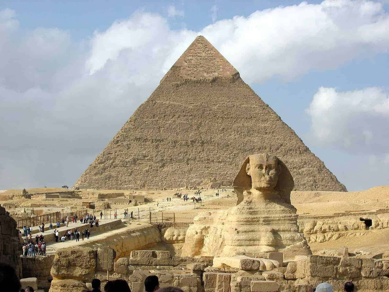 Ancient Egypt 桌布 壁紙 — 大獅身人面像守護著大金字塔的底部，天空部分多雲，遊客正在探索這片古老的沙漠遺址。