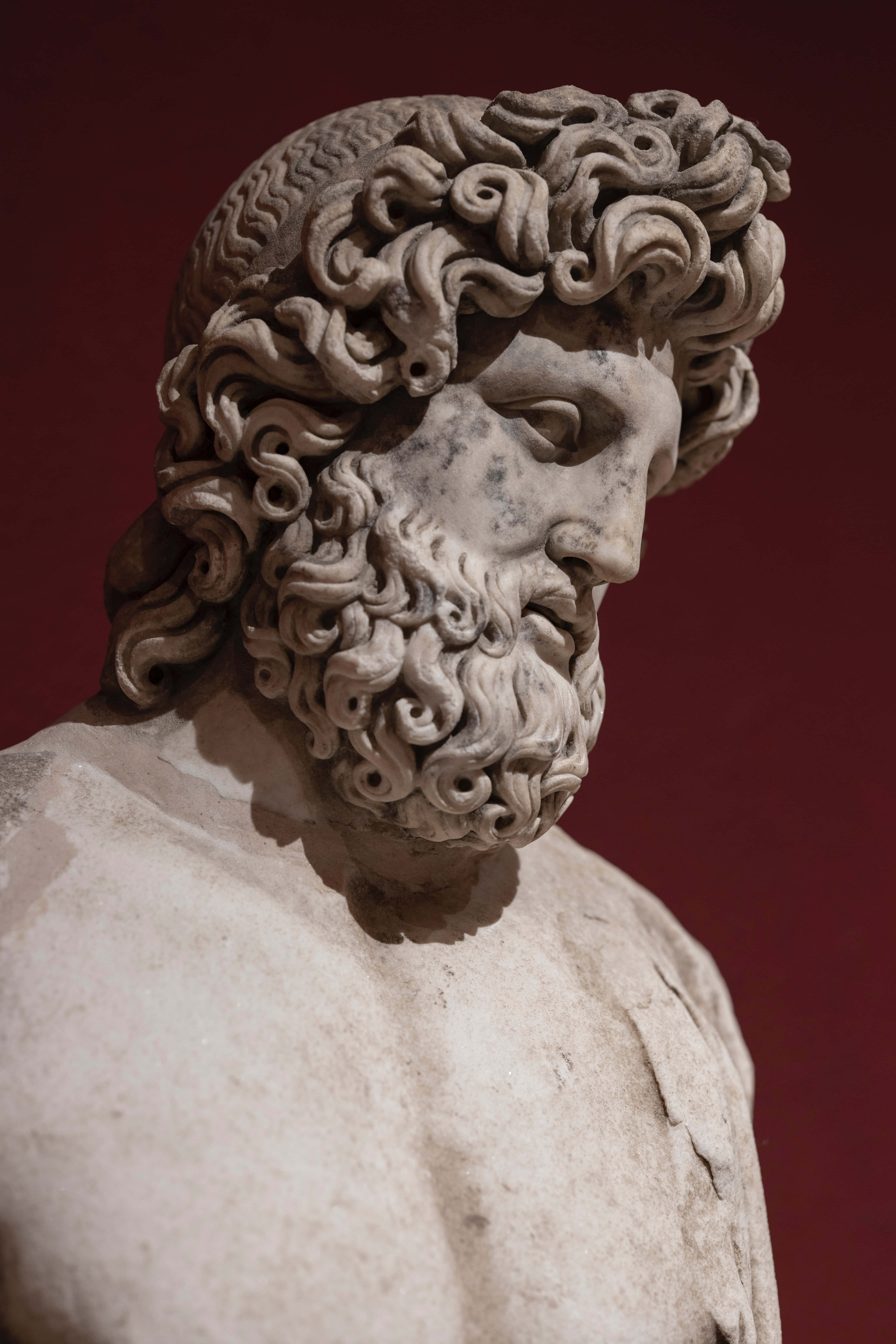 Best Greek Mythology 桌布 壁紙 — 大理石雕像頭部特寫，帶有複雜的捲曲鬍鬚和頭髮，背景為深紅色。