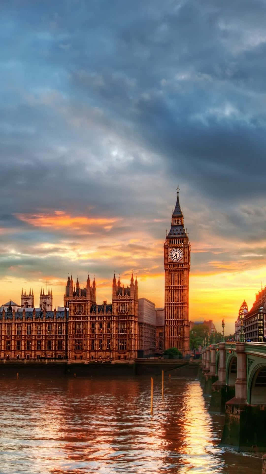 London iPhone 桌布 壁紙 — 大笨鐘和國會大廈在日落時溫暖地發光，它們的倒影在戲劇性的多雲天空下在泰晤士河上閃爍。