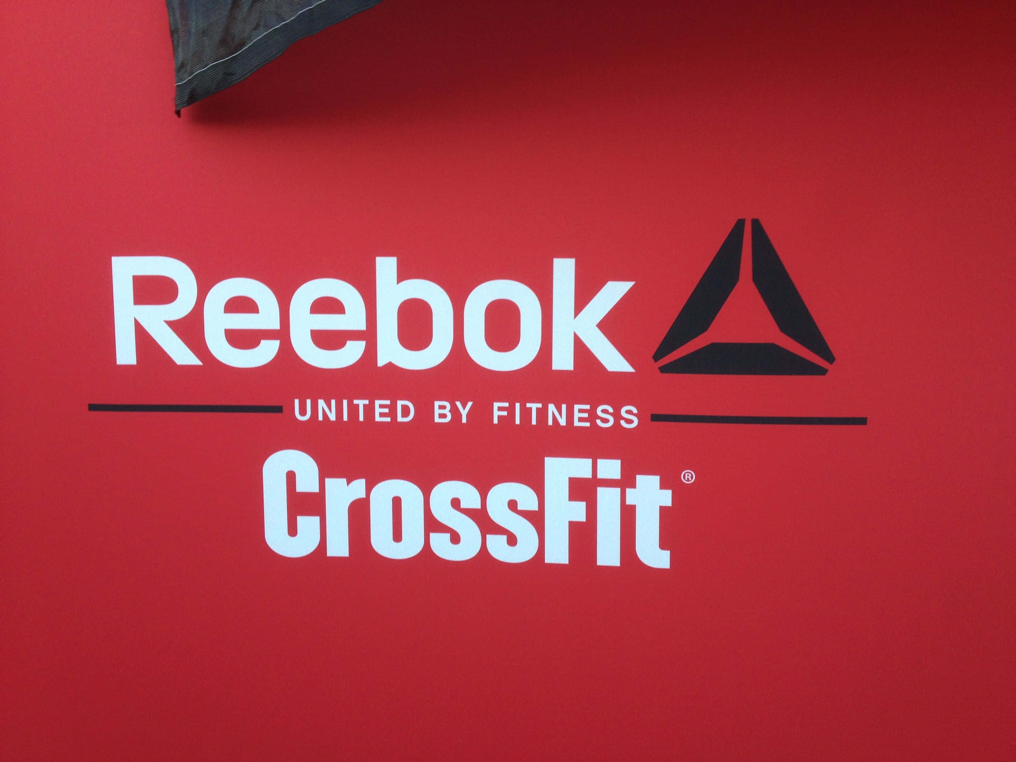 銳步 桌布 壁紙 — 大膽的白色和黑色Reebok CrossFit標誌在充滿活力的紅色牆上，上方有一個深色布料角落。