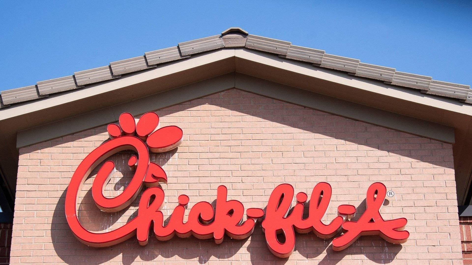 chick 桌布 壁紙 — 大膽的紅色Chick-fil-A標誌安裝在磚砌建築上，背景是晴朗的藍天。