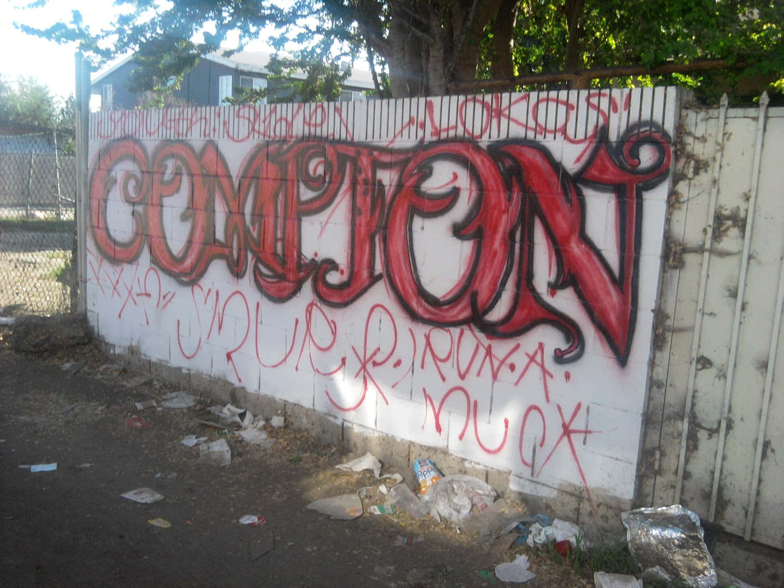 Sur 13 桌布 壁紙 — 大膽的紅色塗鴉拼出「COMPTON」，在一個陽光普照、垃圾遍地的城市小巷中，主宰著一道風化的白色籬笆。