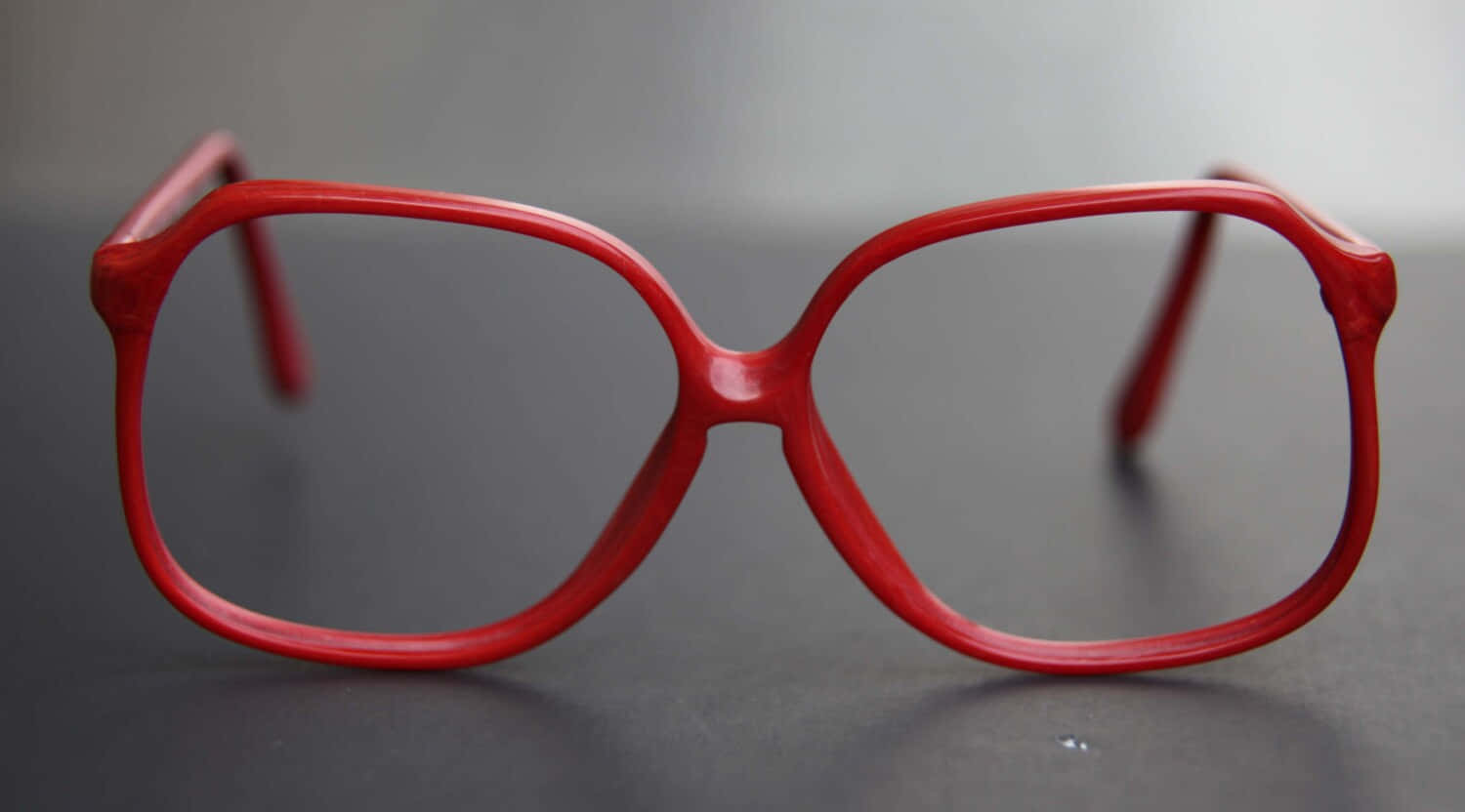 glasses 桌布 壁紙 — 大膽的紅色復古眼鏡，厚框，放在深色反光表面上，背景為柔和的灰色。