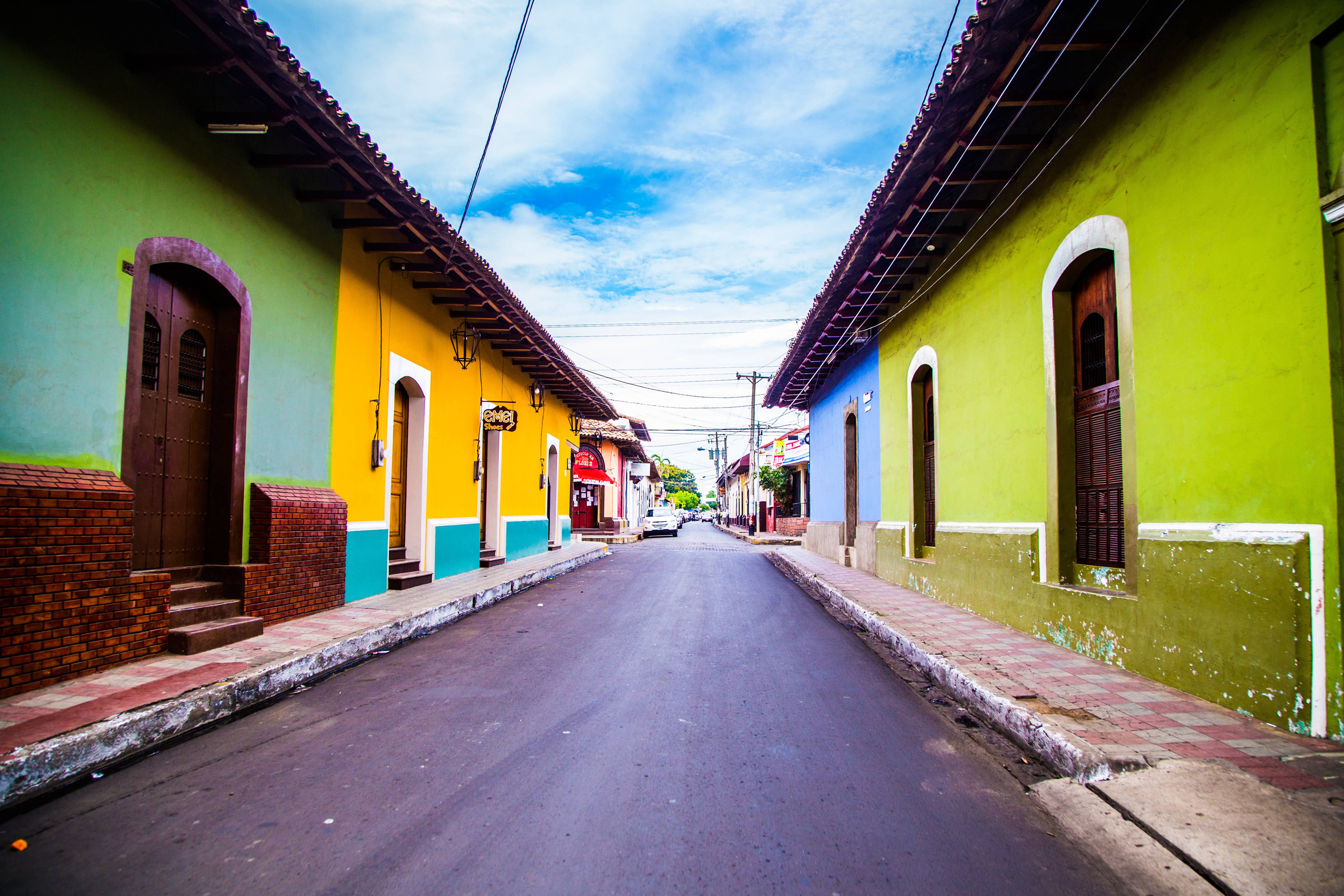 Nicaragua 桌布 壁紙 — 大膽的綠色、黃色和藍色的鮮豔殖民建築排列在一條寧靜的街道上，上方是帶有零星雲朵的明亮藍天。