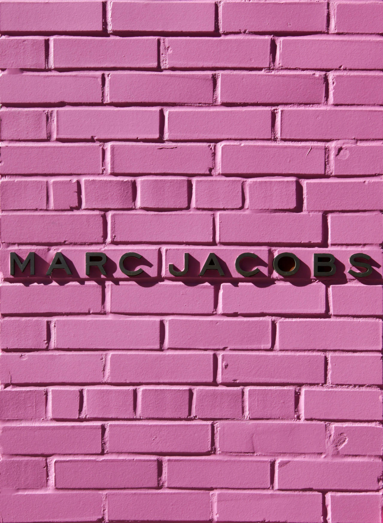 馬克·雅可布 桌布 壁紙 — 大膽的黑色 'MARC JACOBS' 字母安裝在充滿活力的粉紅色油漆磚牆上。