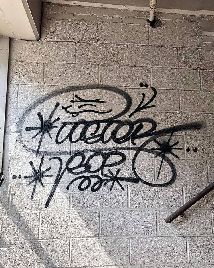 Black And White Graffiti 桌布 壁紙 — 大膽的黑色塗鴉標籤，帶有星爆圖案，噴塗在屋簷下有紋理的白色磚牆上。