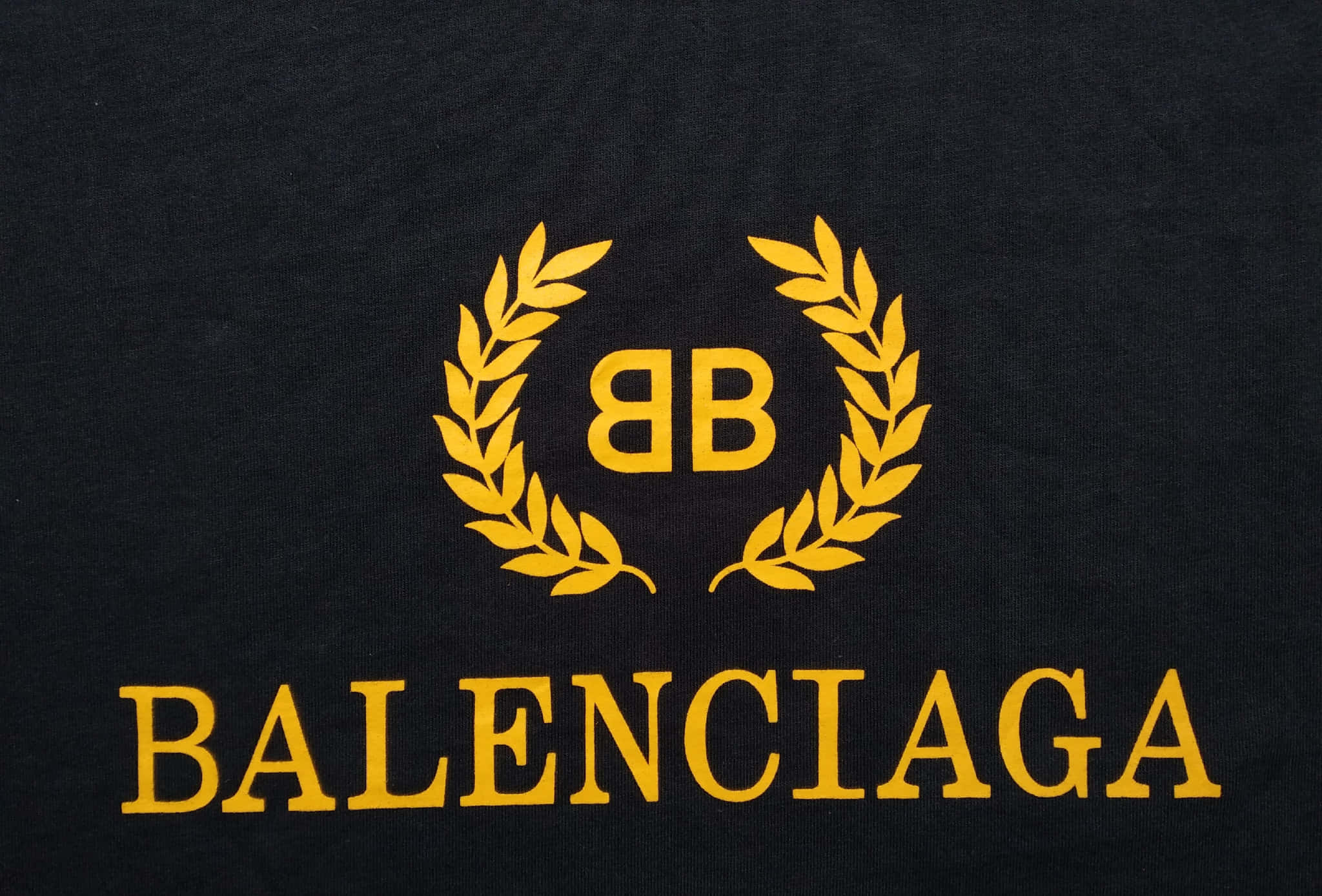 巴黎世家 桌布 壁紙 — 大膽金色 Balenciaga 標誌配月桂花環，特寫於紋理黑色布料上。