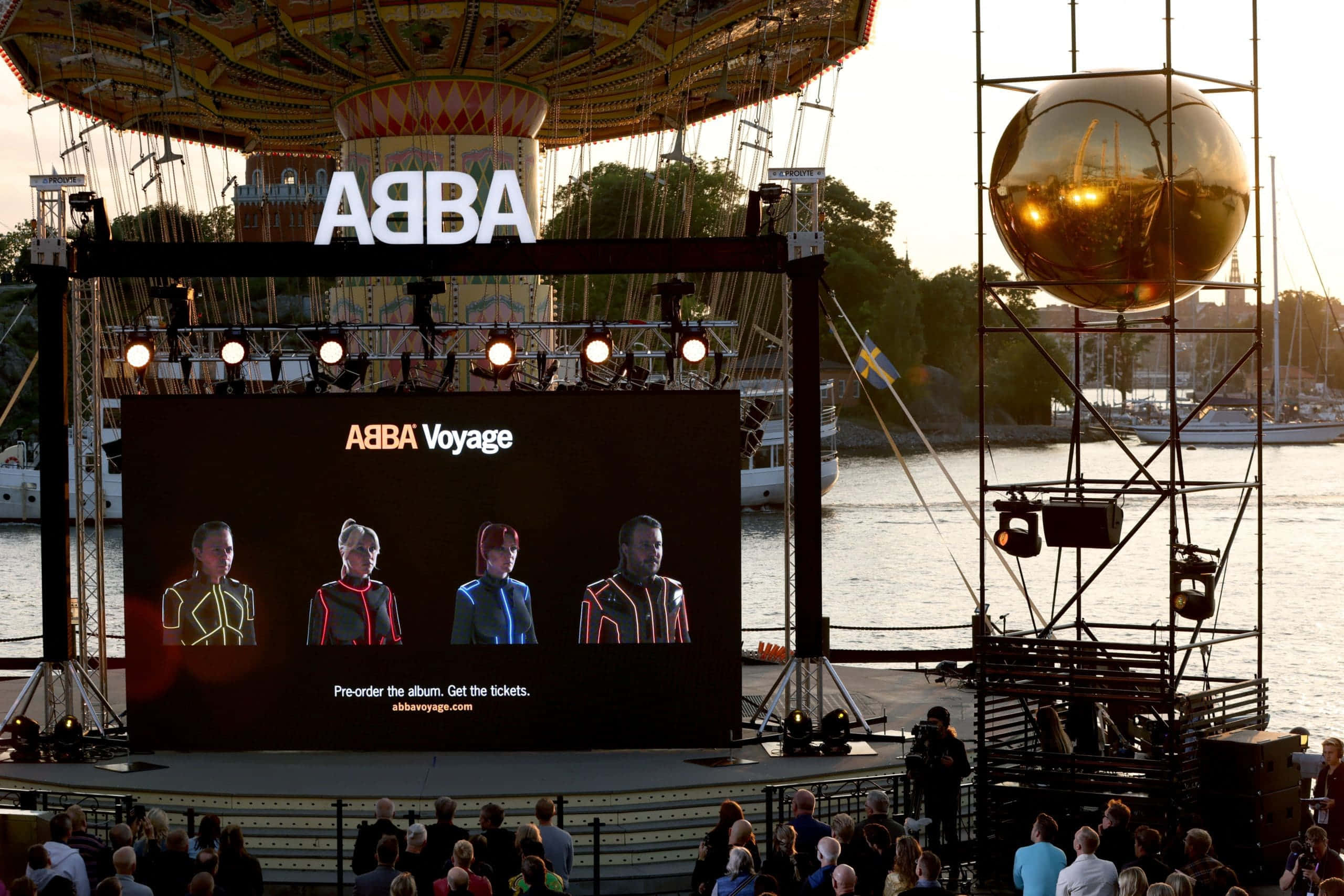 阿巴 桌布 壁紙 — 大螢幕顯示ABBA Voyage，四位表演者穿著未來主義服裝，在日落時分的海濱舞台上演出。